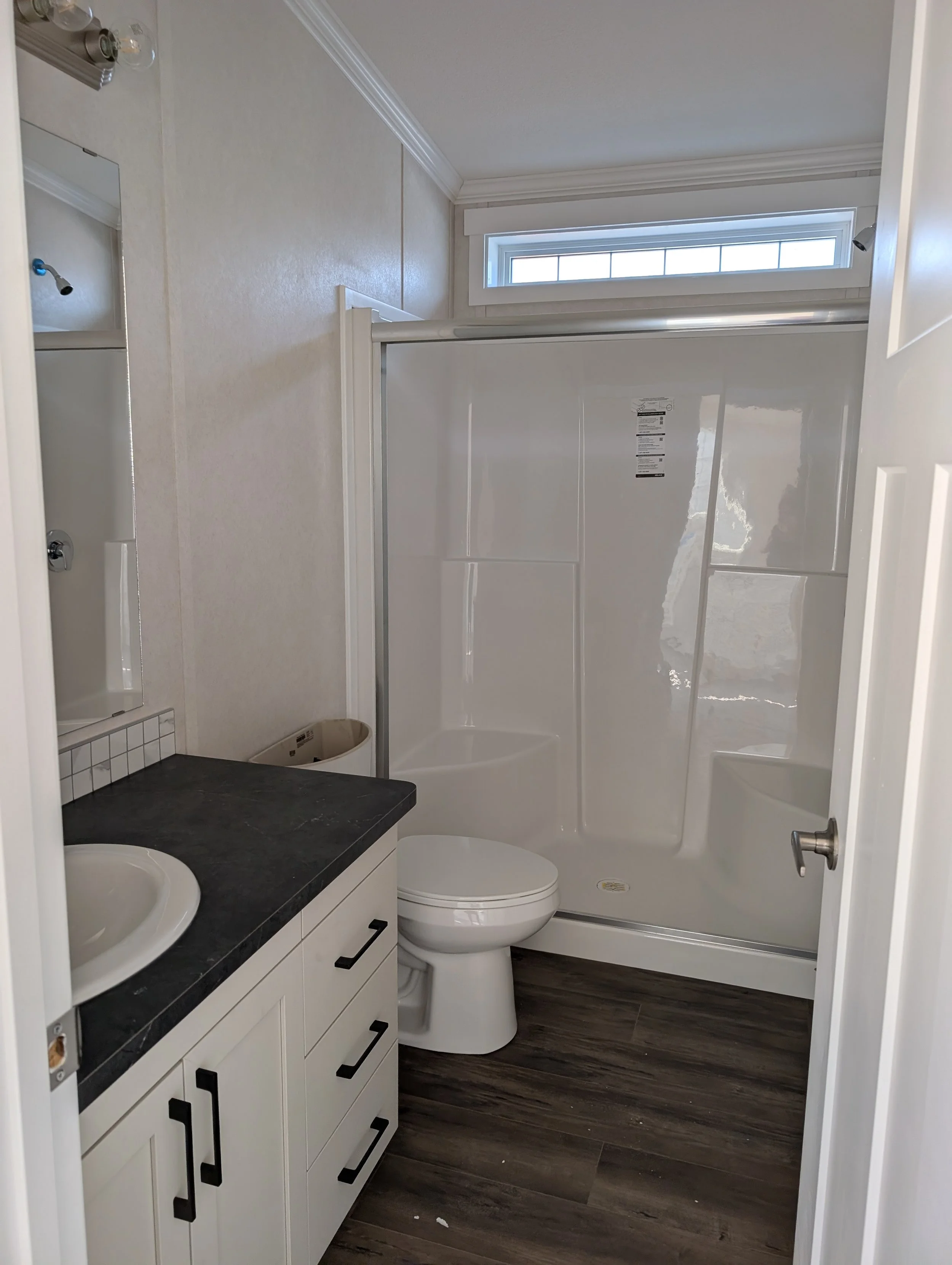 Small bathroom featuring a shower stall with a clear glass door, a white toilet, a black granite countertop with two sinks, and a large mirror above the sinks. The room has a window near the ceiling, crown molding, and wood-style flooring.