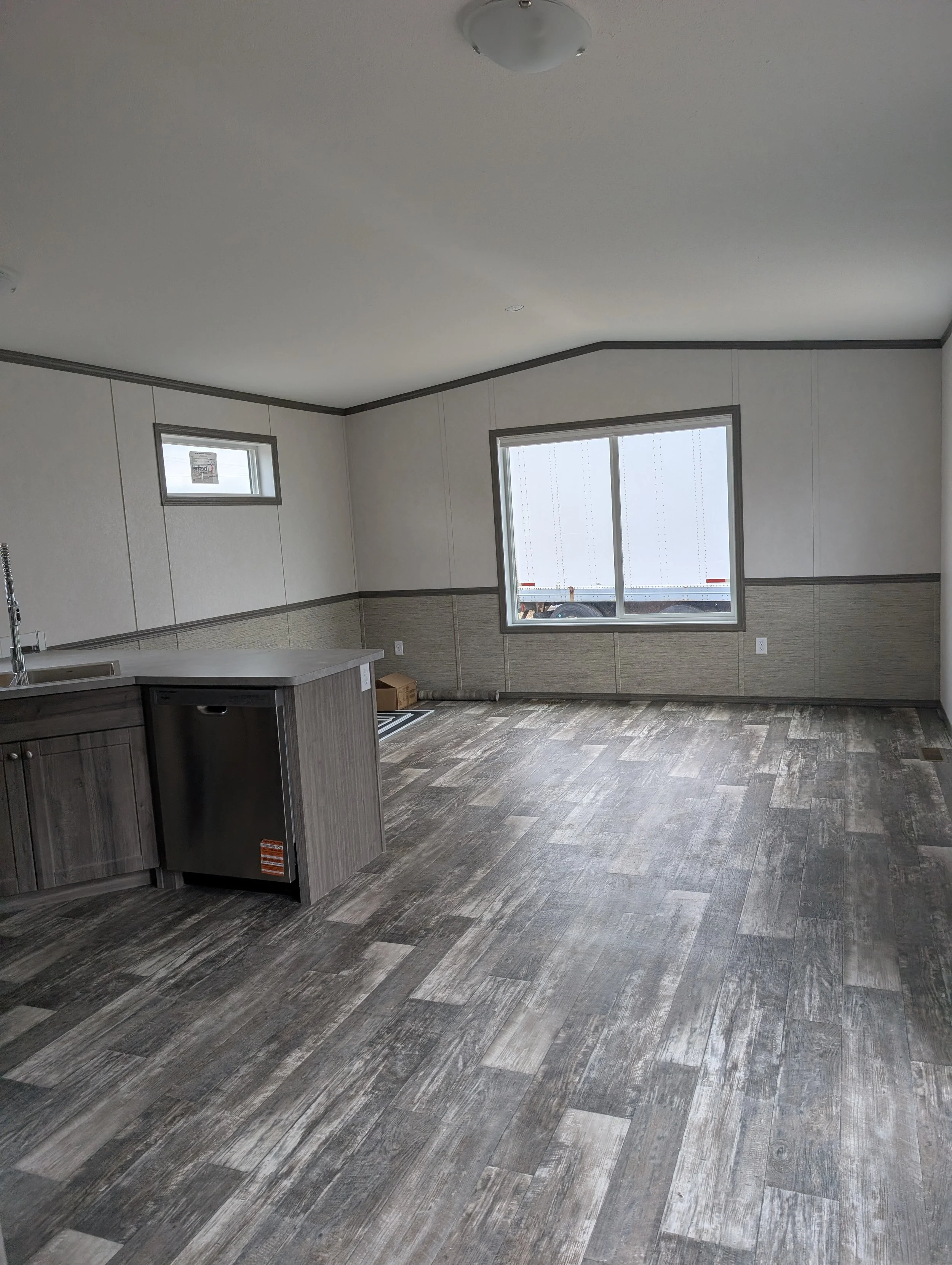 Empty kitchen or dining area with gray wood-like flooring, white walls, gray trim, large window, smaller window, and a kitchen counter with sink in the corner.