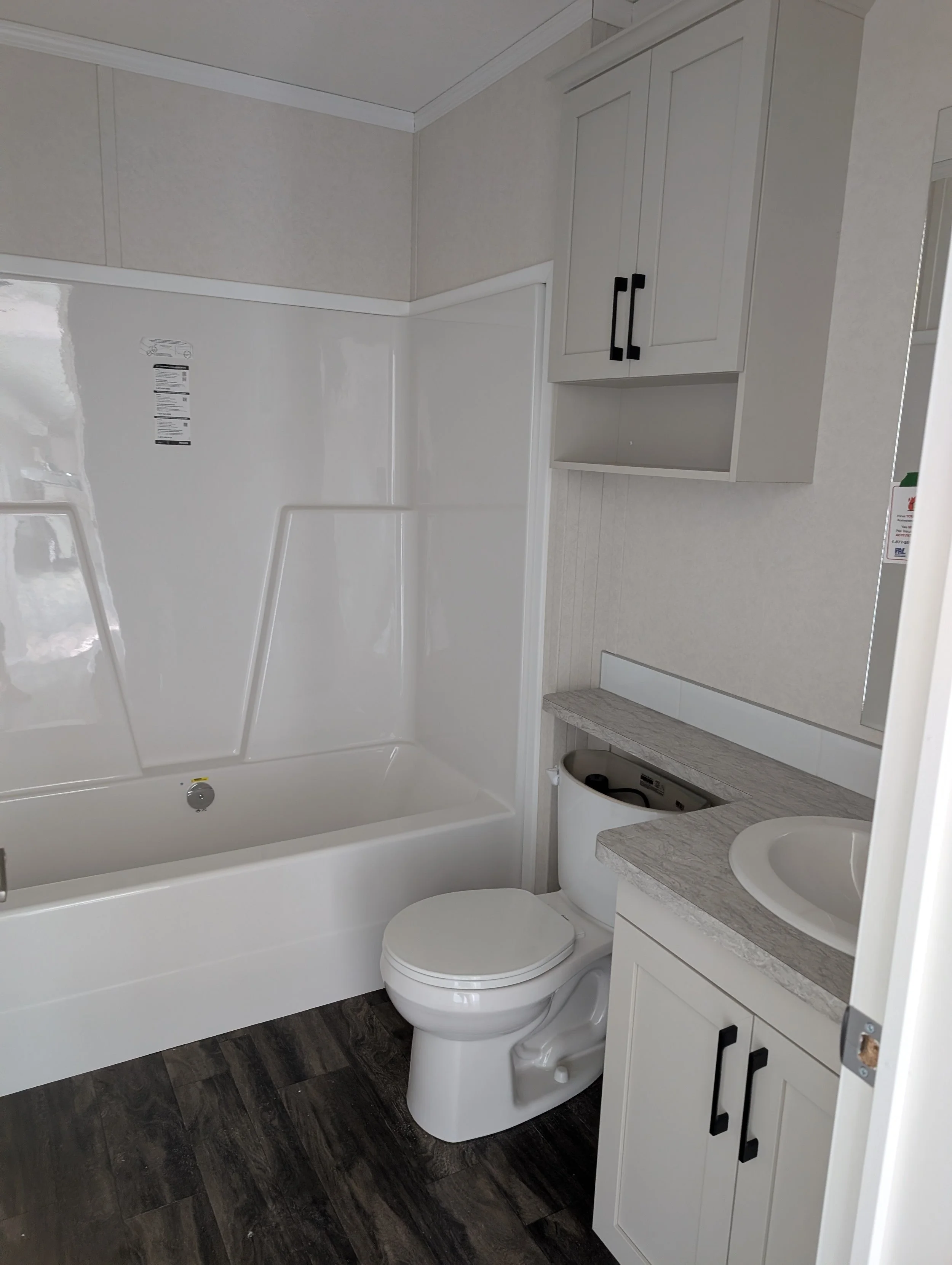 Bathroom with a bathtub, a toilet, a white vanity with a marble countertop, and a white cabinet with black handles. Dark wood-look flooring and cream-colored walls.