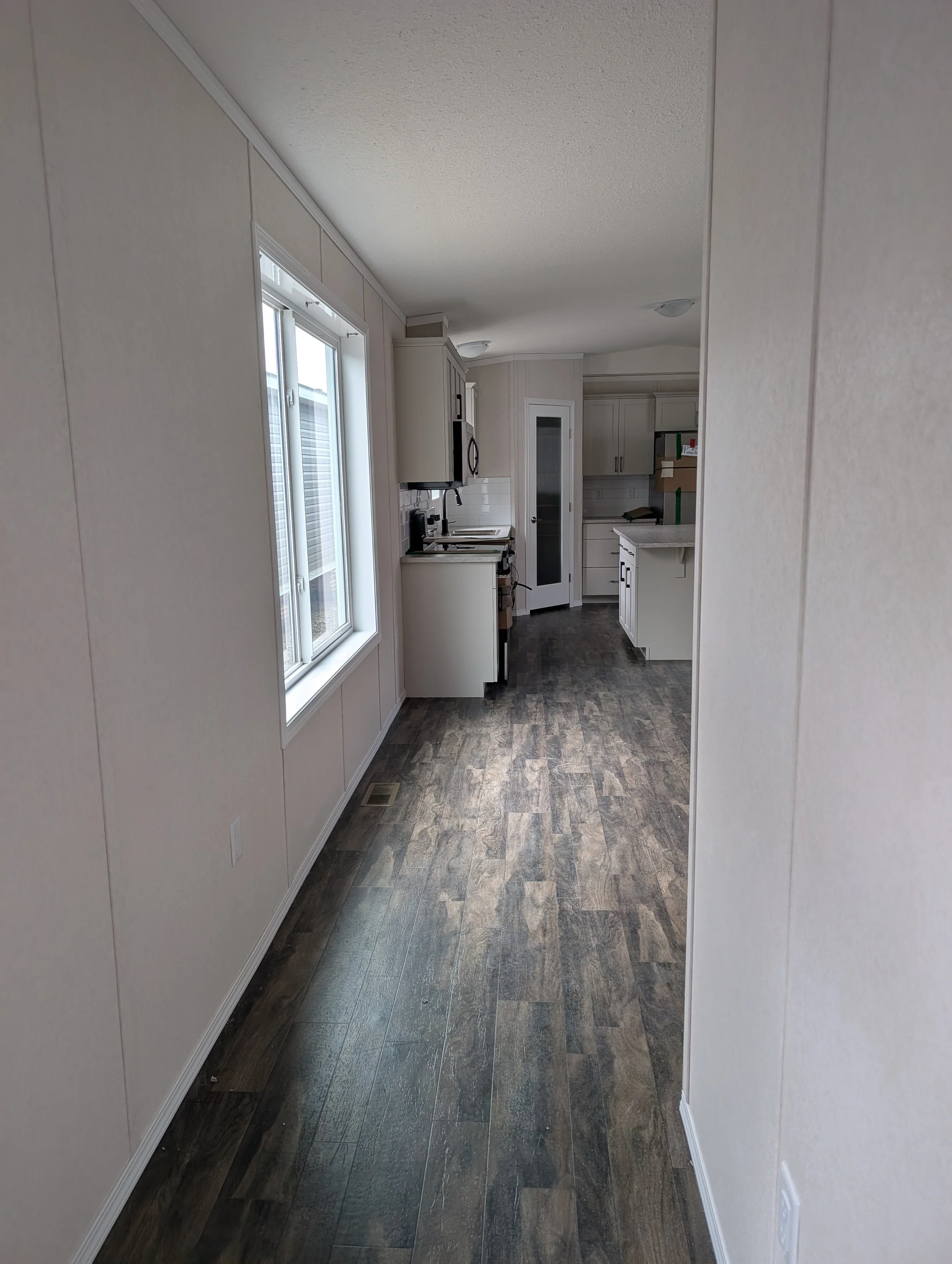 A hallway inside a house with beige walls, dark wood flooring, a large window on the left, and white kitchen cabinets visible in the background.