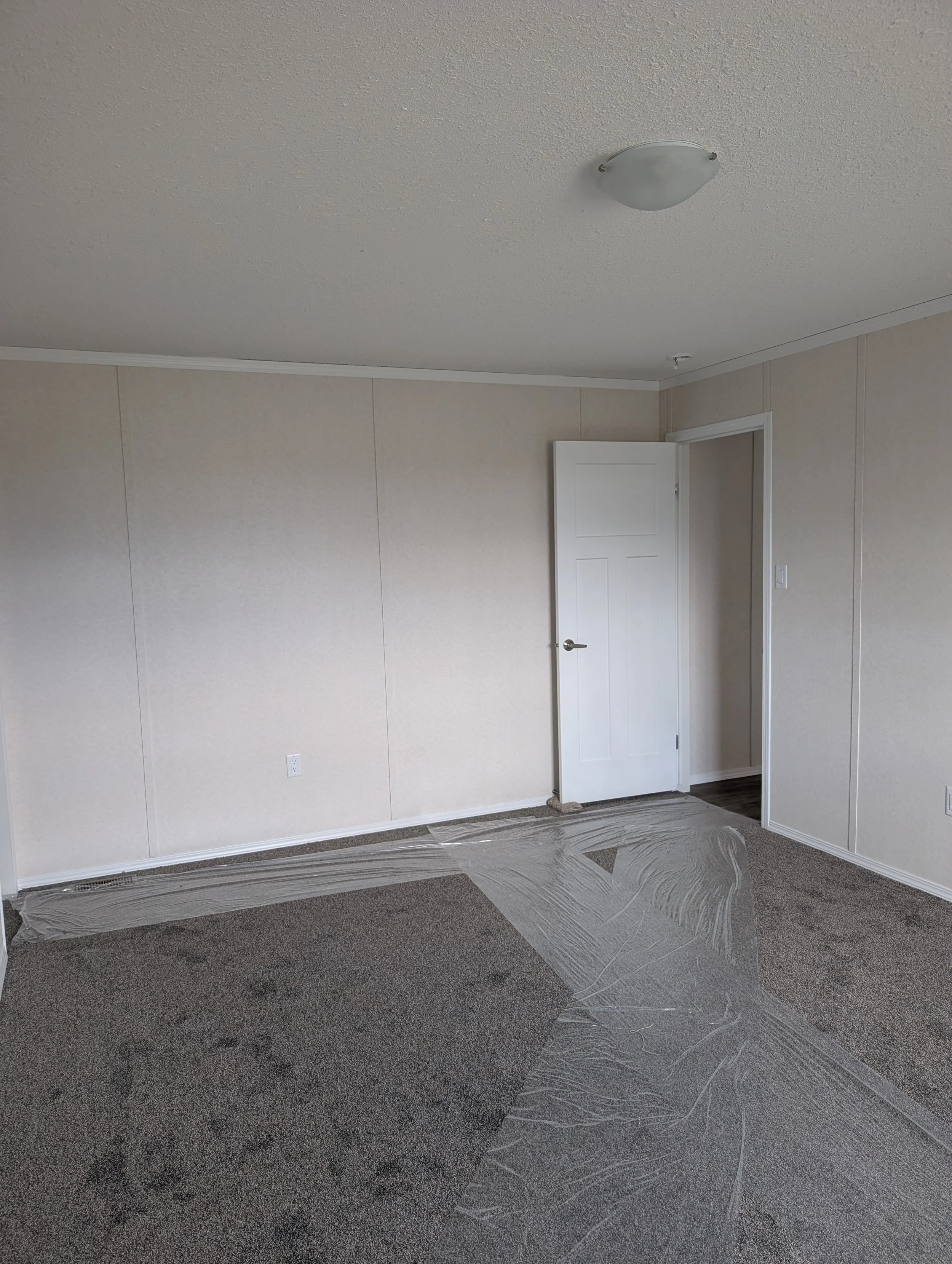 Empty room with beige walls, a white door slightly open, carpeted flooring, and plastic protective covering on the floor.