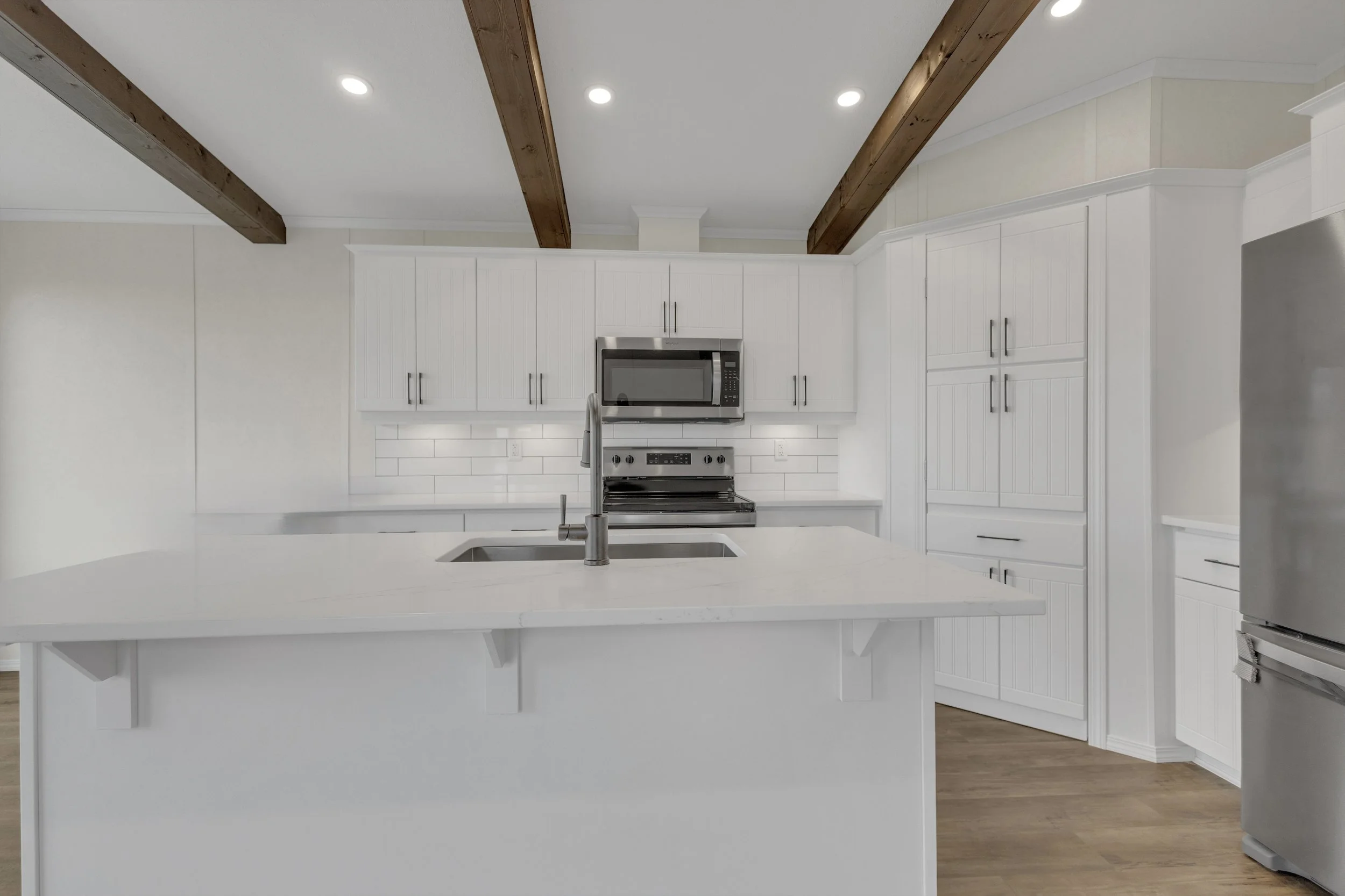 Modern white kitchen with stainless steel appliances, white cabinets, a white countertop island with a sink, and wooden beams on the ceiling.
