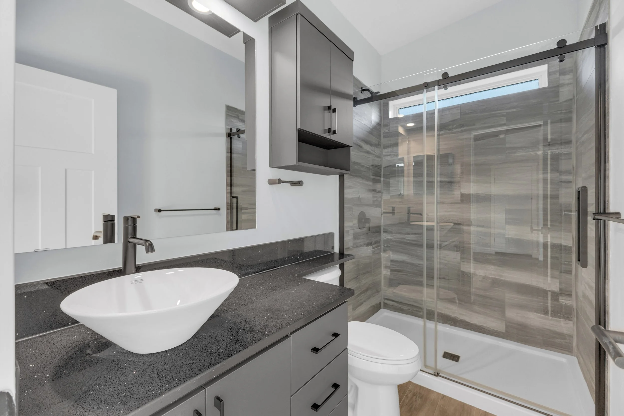 Modern bathroom with gray cabinetry, countertop with a vessel sink, wall-mounted mirror, glass shower enclosure with sliding door, and gray stone wall tiles.