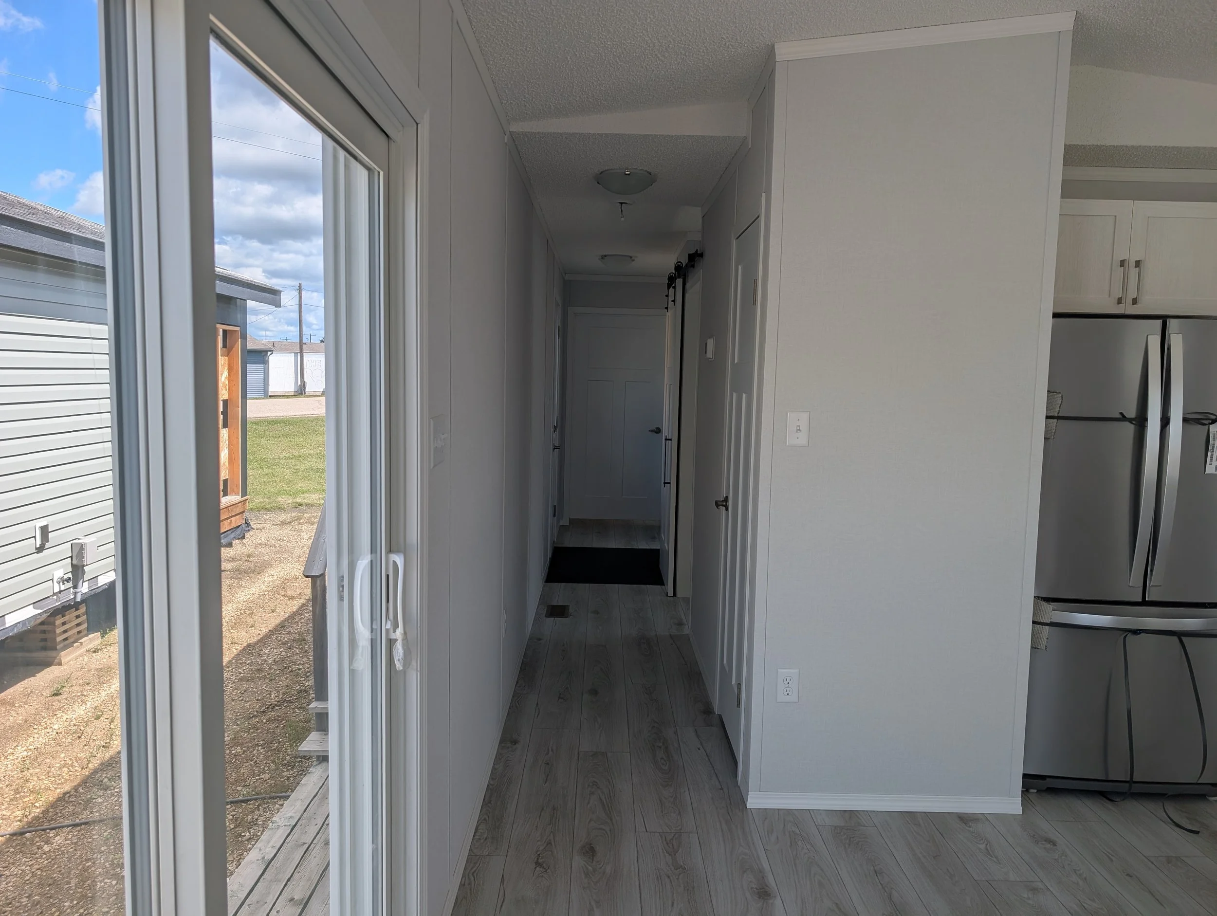 Inside view of a kitchen with white cabinets, a gray refrigerator, and large windows looking outside to a grassy yard with a building and cloudy sky.