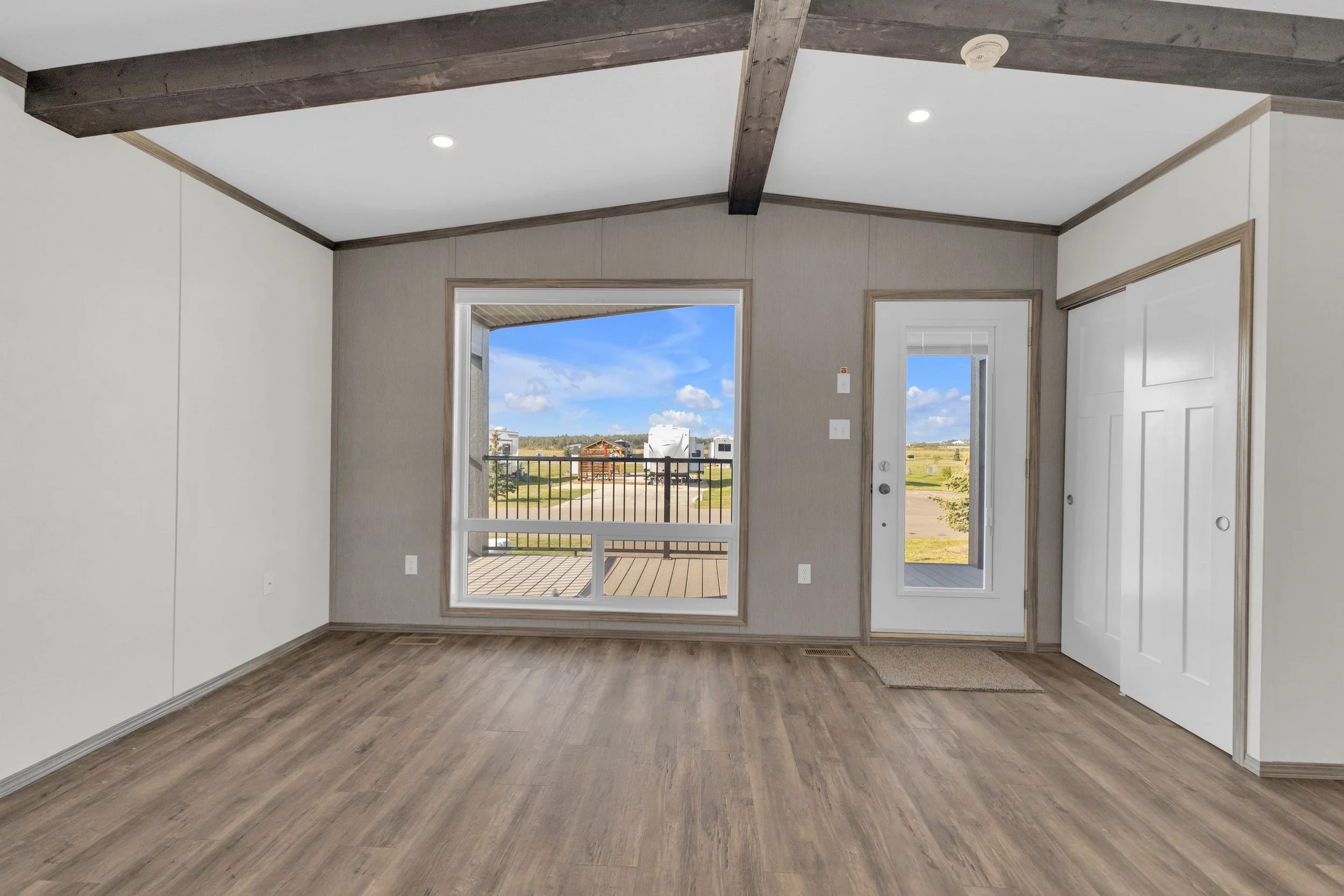 Empty room with wood flooring, white walls with wood trim, large window and glass door leading to a balcony, with a view of a blue sky and fields outside.