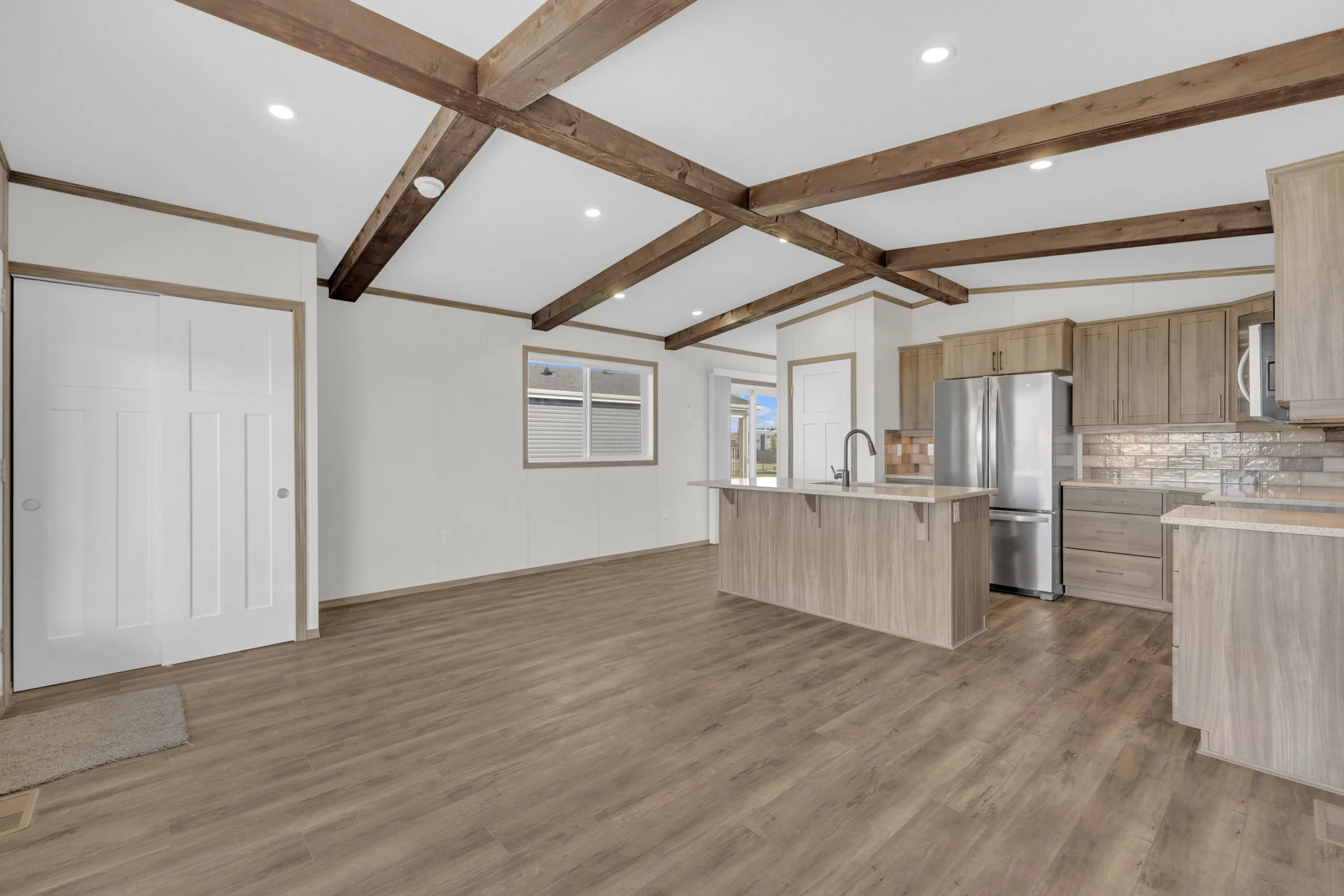 A modern kitchen with light wood cabinets, a stainless steel refrigerator, and a kitchen island. The ceiling has exposed wooden beams and recessed lighting. There are two windows, and a door leading outside.