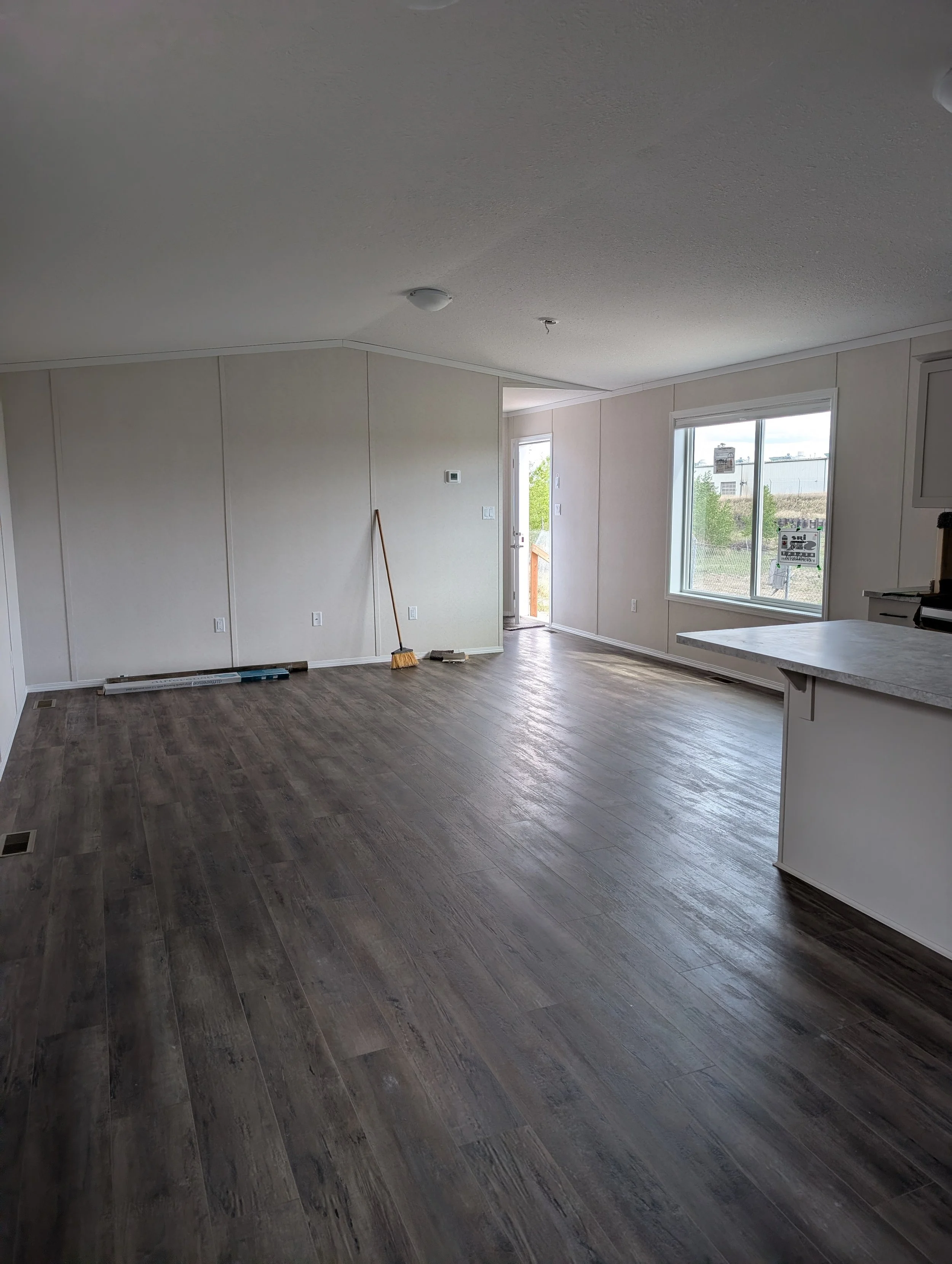 Empty living room with wood flooring, large window, and front door, with cleaning tools on the floor, in a house under construction or renovation.