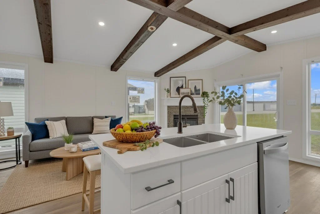 Modern open-concept kitchen and living room with white cabinets, a central island with a sink and a fruit bowl, a gray sofa with throw pillows, and large windows showing a grassy outdoor view.