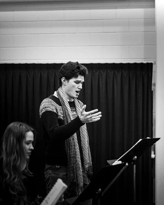 A young man with dark hair, wearing a striped scarf and a sweater, is standing at a music stand, singing or speaking, with a focused expression. A young woman with long hair, holding a book or script, is sitting nearby, listening attentively. The scene is in a room with a black curtain and white brick wall in the background.