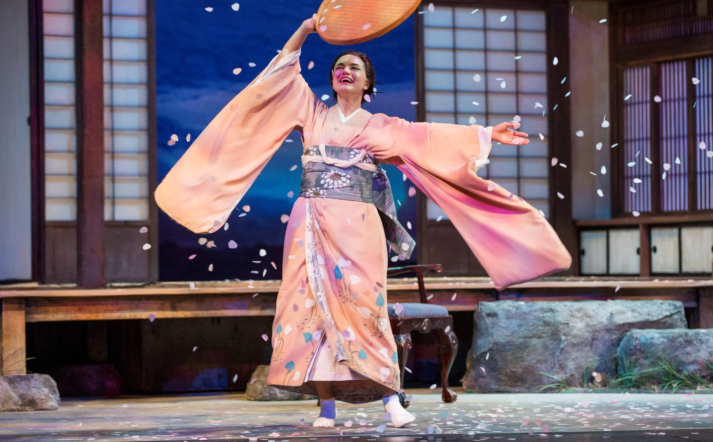 A woman in traditional Japanese kimono dancing on stage with cherry blossom petals falling around her, in front of a backdrop resembling a Japanese room.