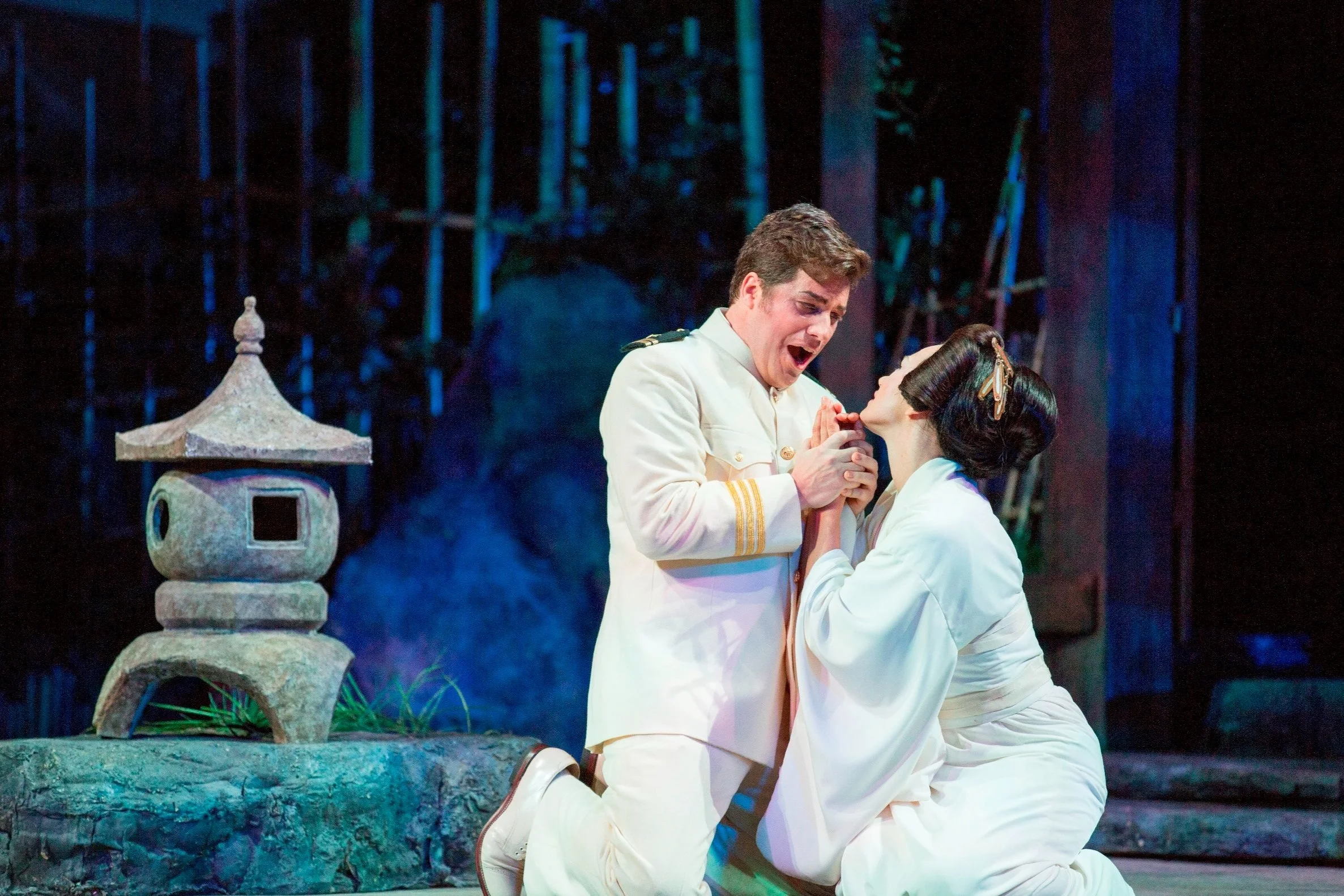 Two actors in a theatrical production, dressed in white, are engaging in an emotional scene on stage. The man is kneeling and appears distressed, while the woman is kneeling as well, holding his hands and looking up at him. The background features a dark, forest-like setting with bamboo and a traditional stone lantern.