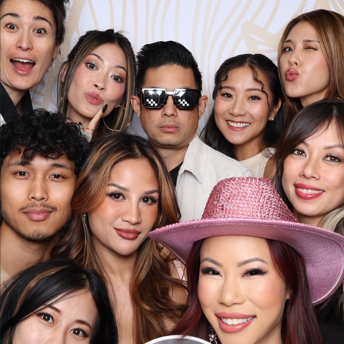 Group of diverse young adults taking a selfie, with one woman wearing a pink hat, in a party setting.