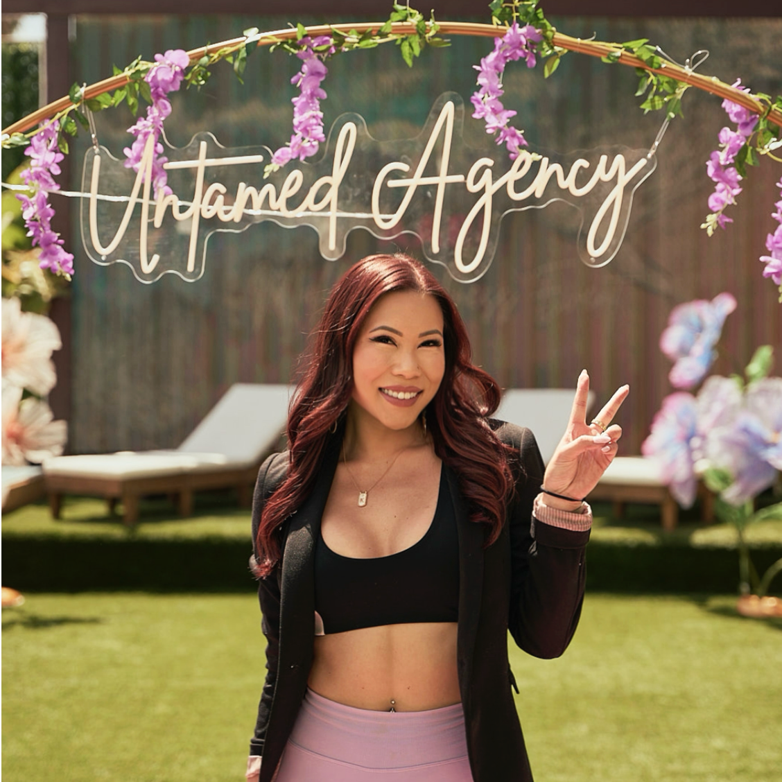 A woman smiling and making a peace sign at an outdoor event, standing in front of a sign that reads 'Untamed Agency', decorated with purple flowers.