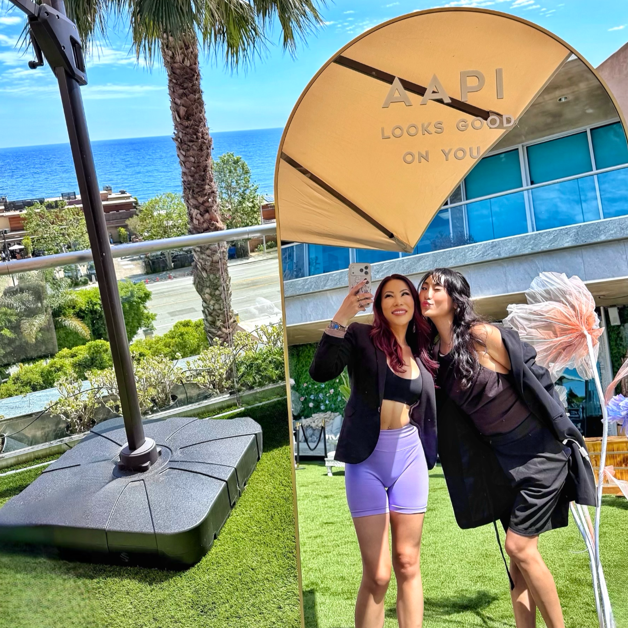 Two women taking a selfie under a large beige umbrella that has the words 'AAPI Looks Good On You' written on it. One woman has long red hair and is wearing a black crop top and lavender shorts. The other woman has long black hair and is wearing a black blazer and dress. They are smiling and standing on a grassy area with a view of the ocean, palm trees, and a building with large blue windows in the background.