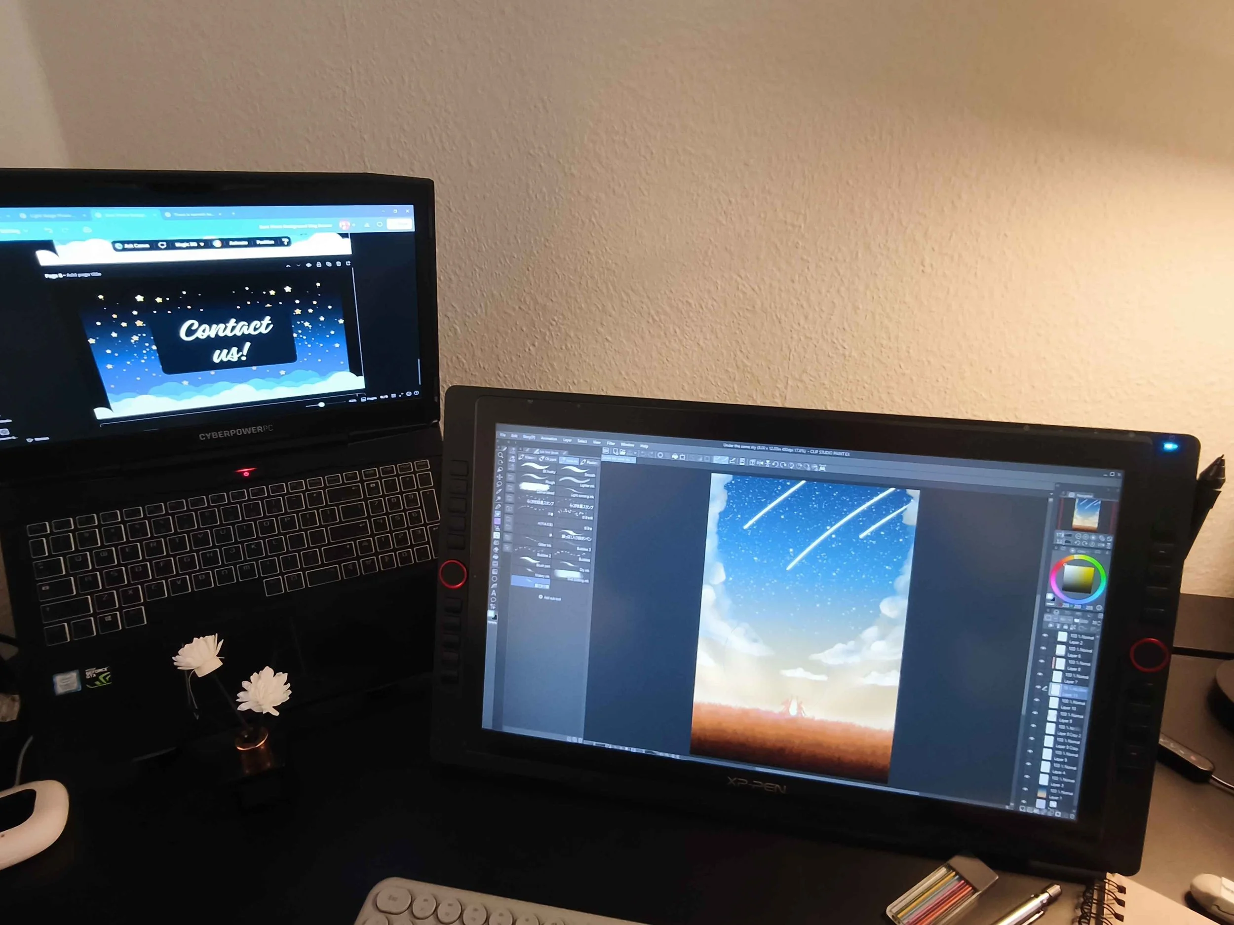 Two computer screens on a desk, one displaying a rock climber on a scenic background, the other showing a digital painting of shooting stars over a landscape. A small flower-shaped lamp and various pens are also on the desk.