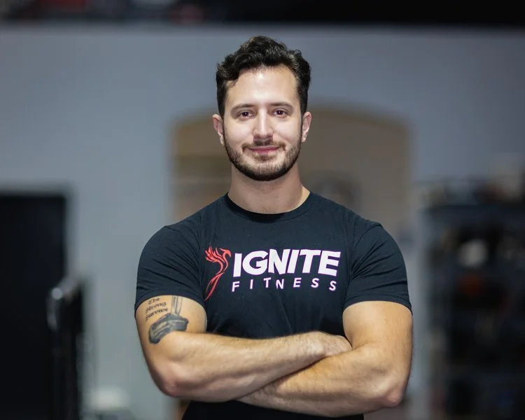 Man with beard smiling, wearing black Ignite Fitness t-shirt, standing with arms crossed in gym environment.