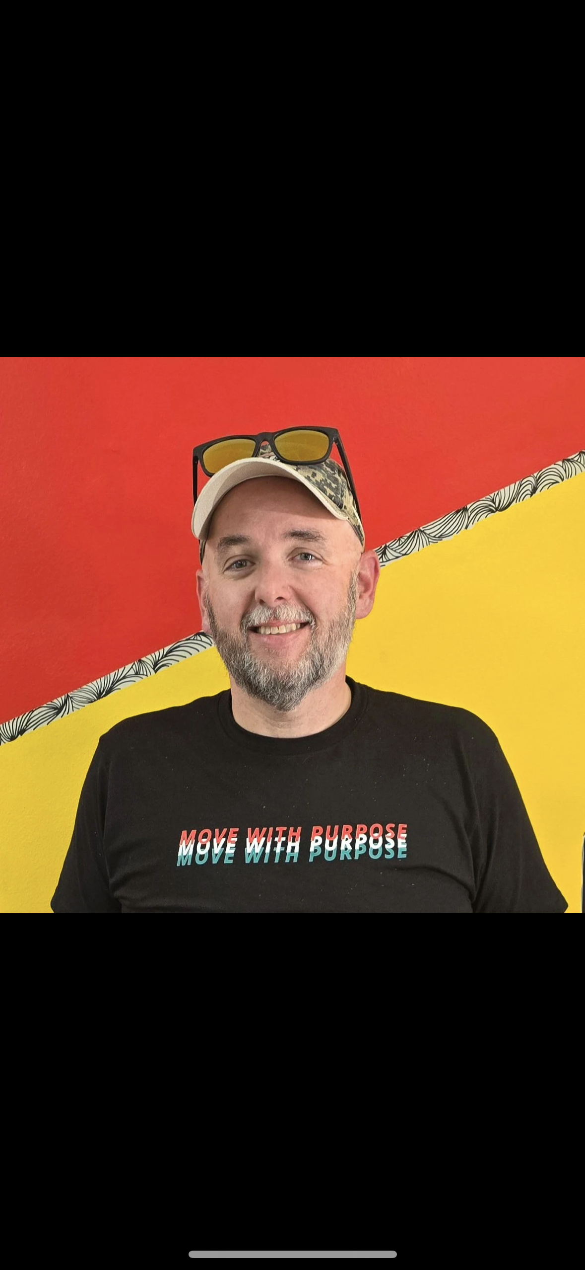Smiling man with a beard and mustache wearing a black 'Move with Purpose' t-shirt, a beige baseball cap, sunglasses on his head, standing against a wall painted with red, yellow, and black and white patterned accent.