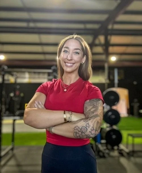 Smiling woman in red shirt with tattooed arms standing in a gym with crossed arms.