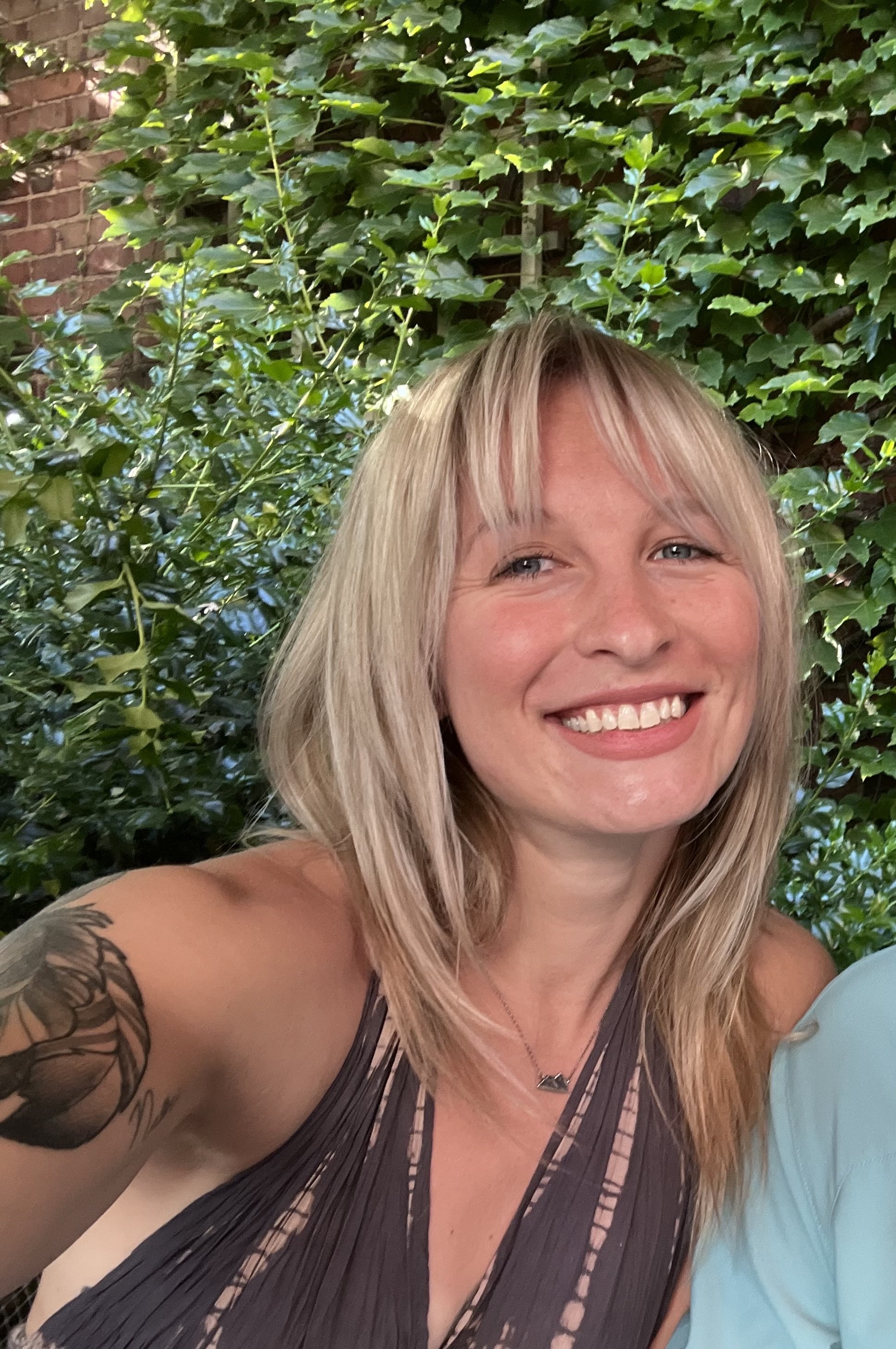 A woman with blonde hair smiling outdoors, surrounded by green ivy and plants.