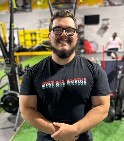 Person smiling, wearing glasses and a "Move with Purpose" shirt, in a gym setting with equipment in the background.