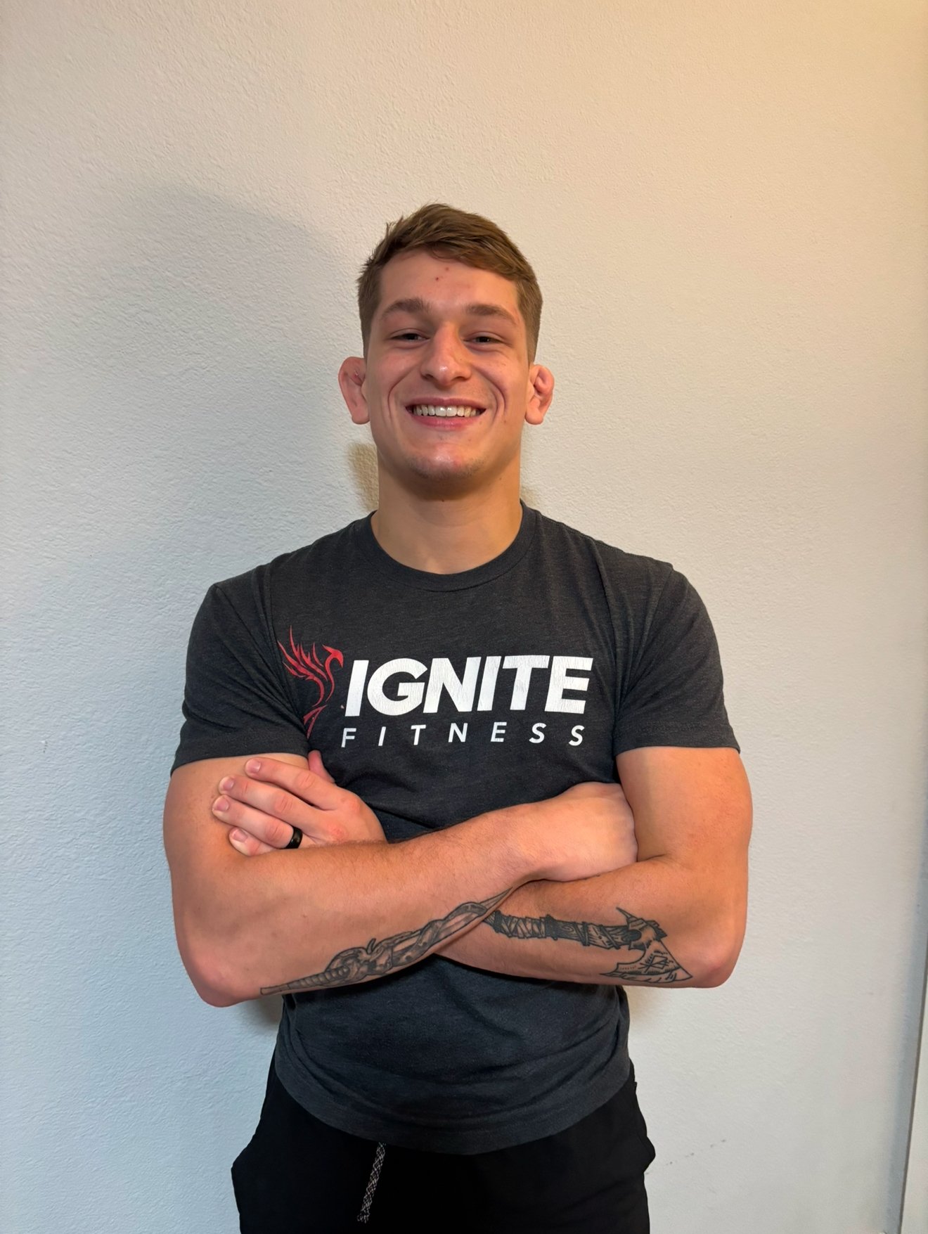 Man smiling in a gray "Ignite Fitness" t-shirt with arms crossed, tattoos visible on arm, against a plain wall.
