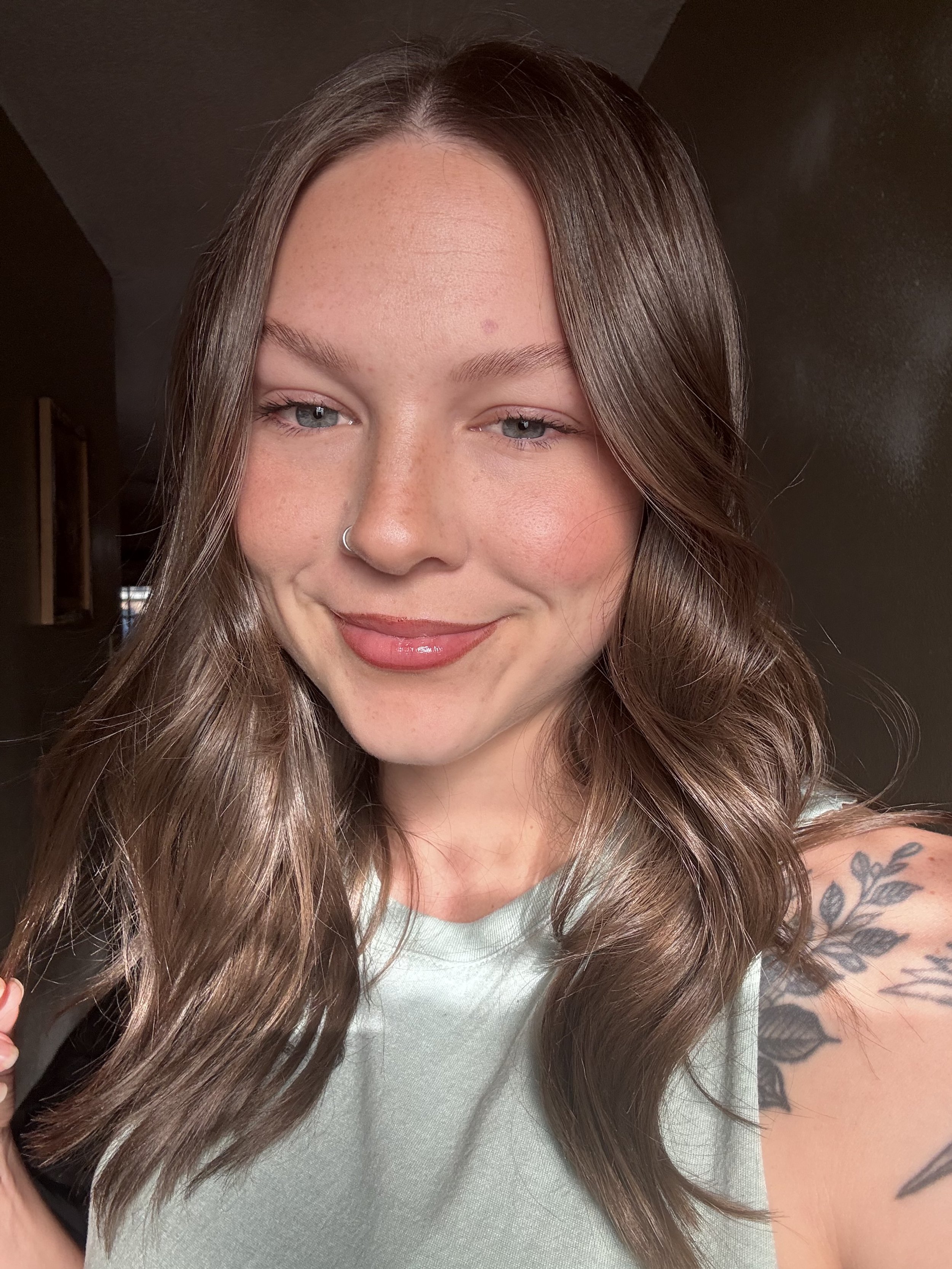 A young woman with long wavy brown hair, wearing a light-colored top, smiling with her eyes closed, and a tattoo of leaves on her shoulder.