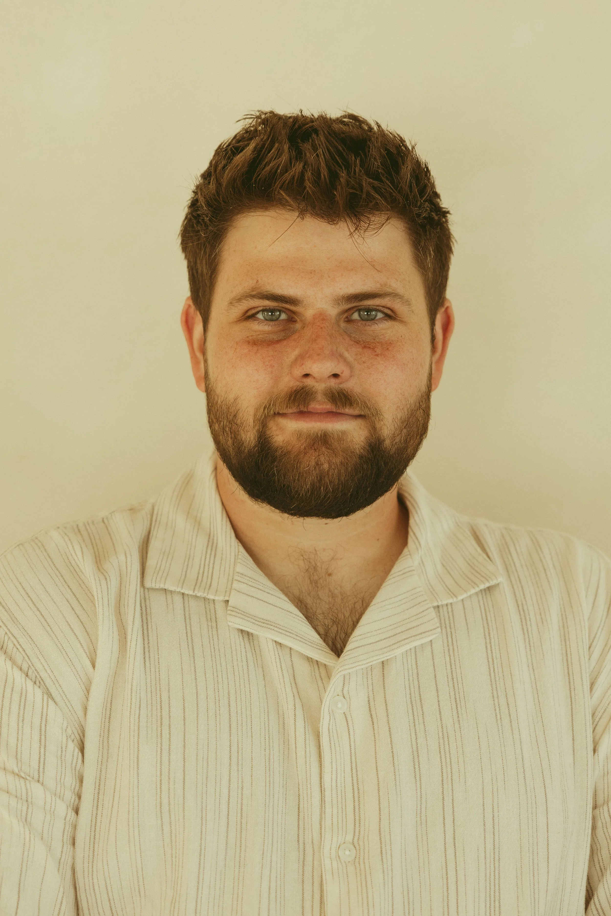 Portrait of a man with short brown hair and a beard, wearing a cream colored shirt, standing against a plain light-colored background.