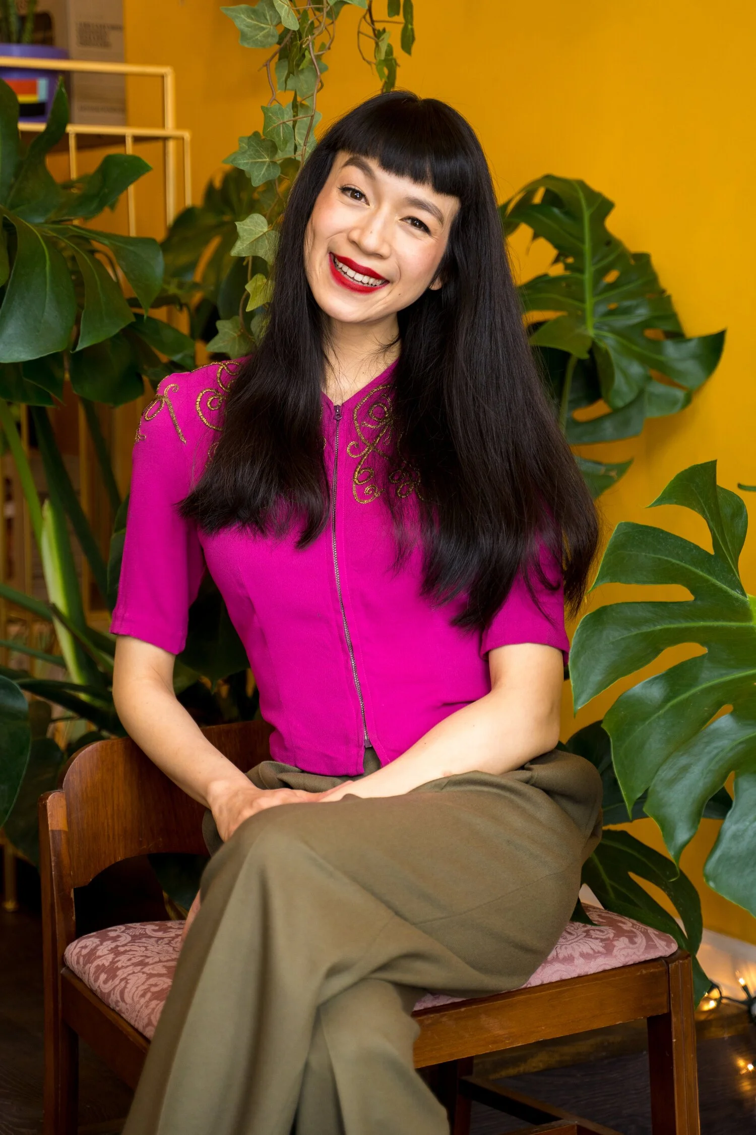 A woman with long black hair, bangs, and red lipstick sitting on a wooden chair with a pink cushion. She is wearing a vibrant pink top with gold embroidery on the shoulder and smiling at the camera. Behind her are large green leaves and a yellow wall.