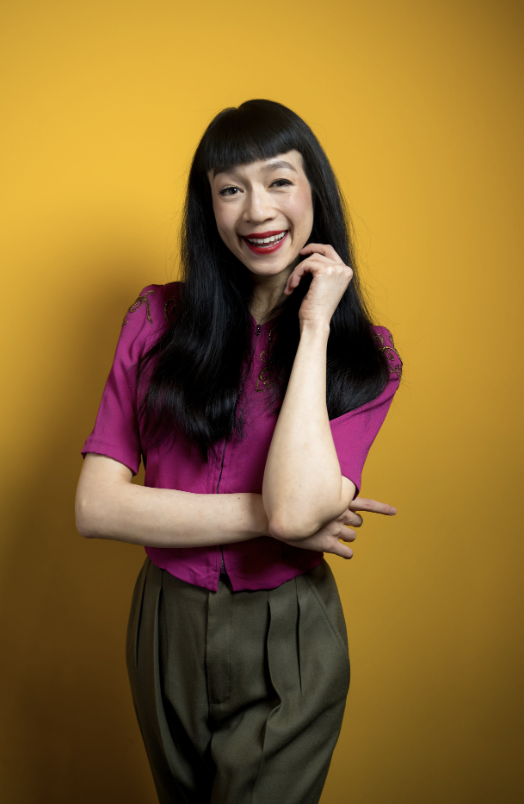 A woman with long black hair, wearing a magenta shirt and khaki pants, smiling and posing with one hand touching her face against a yellow background.