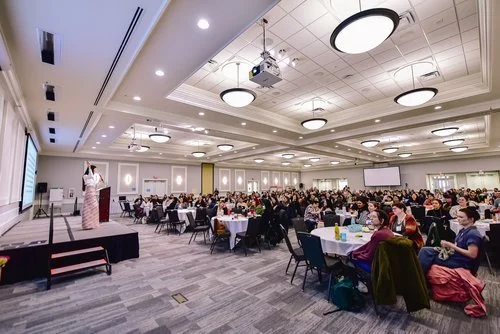 Large conference room filled with seated attendees listening to a speaker on stage with a podium, large screen, and presentation equipment.