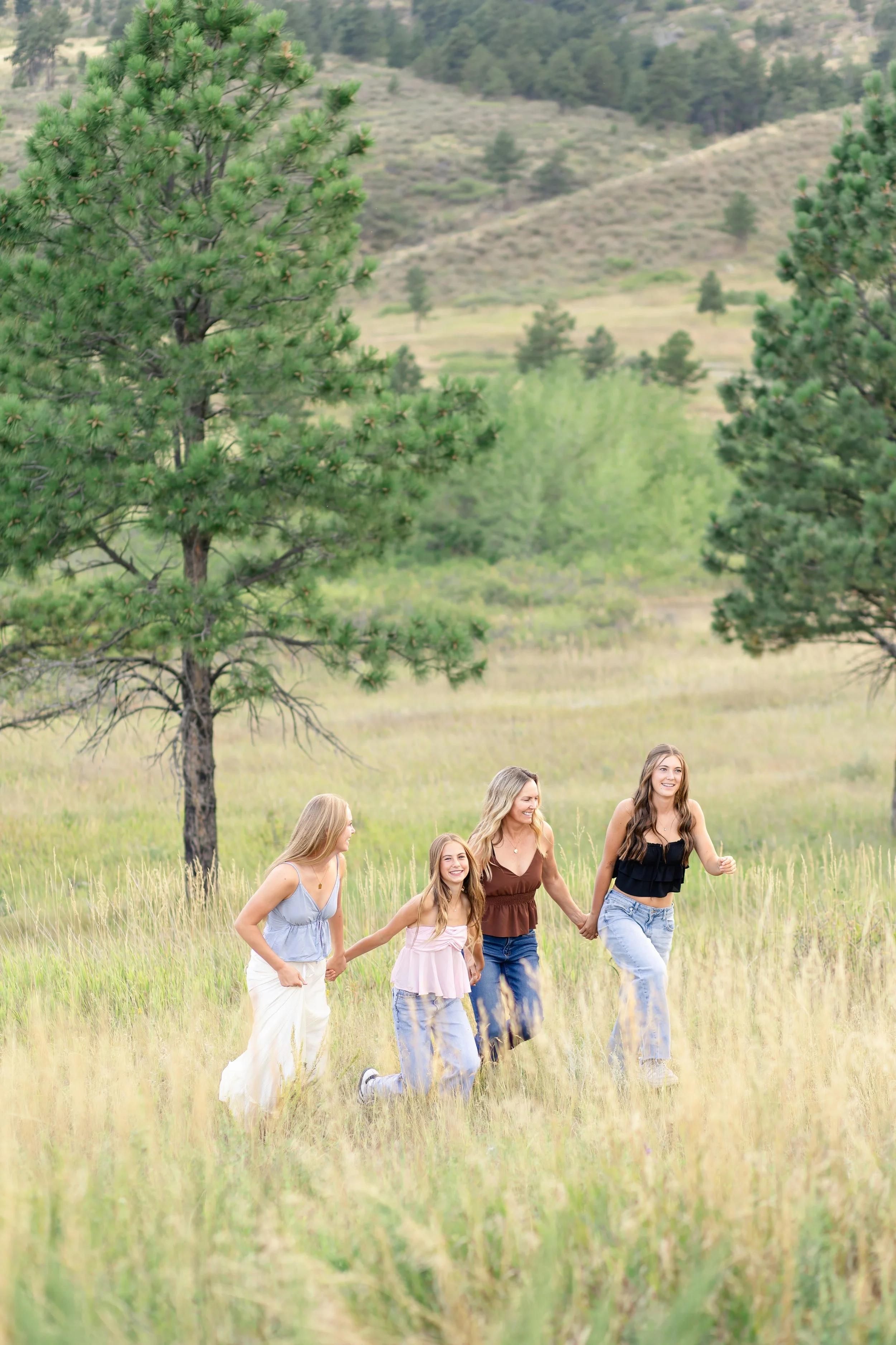 Four women, including two children, walk hand in hand through a grassy field with trees and hills in the background, enjoying a sunny day outdoors.