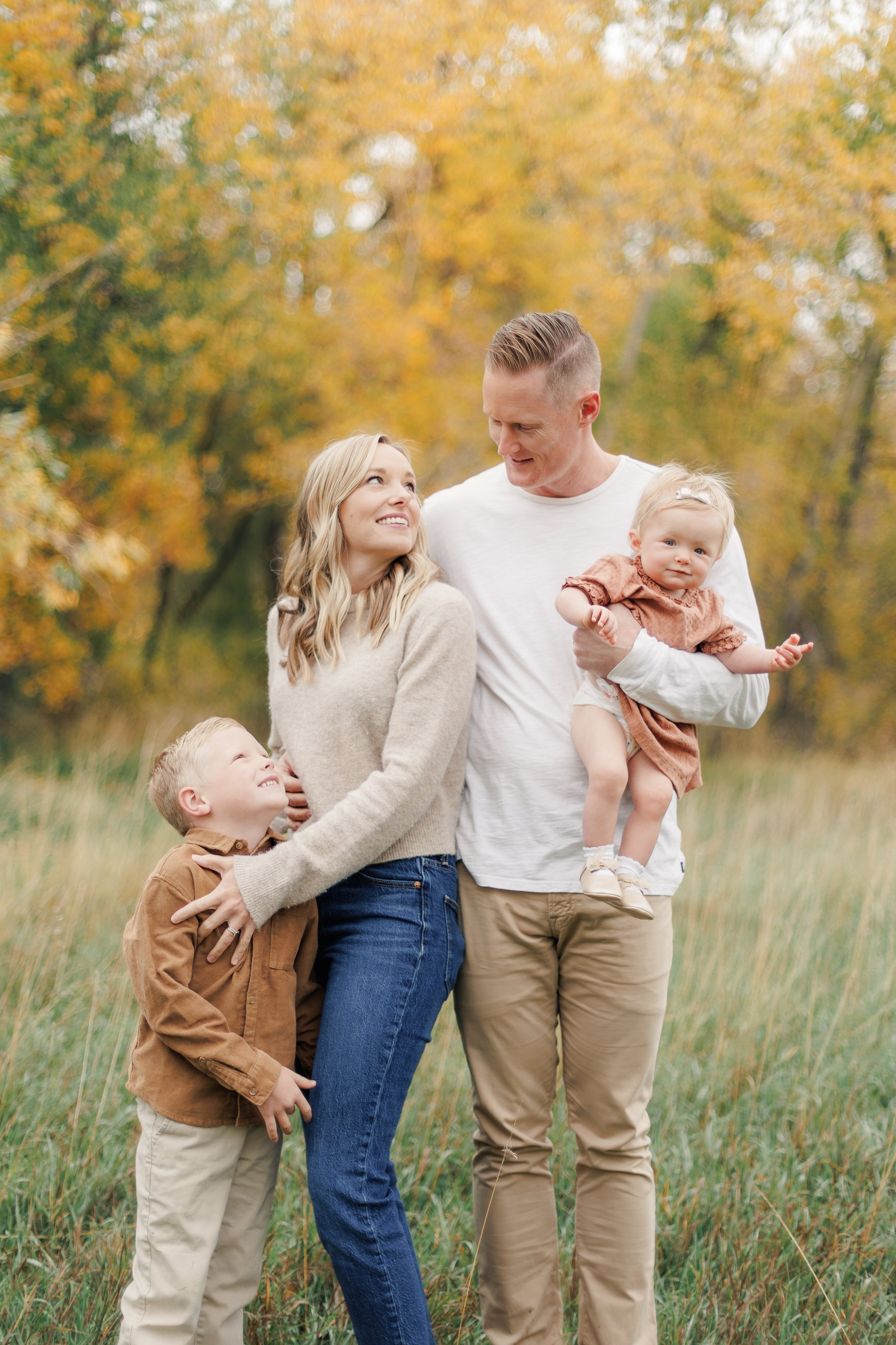 A family of four, including a mother, father, young son, and toddler daughter, standing outdoors in a grassy field with autumn-colored trees in the background. The father is holding the daughter, and the mother is smiling at the son.