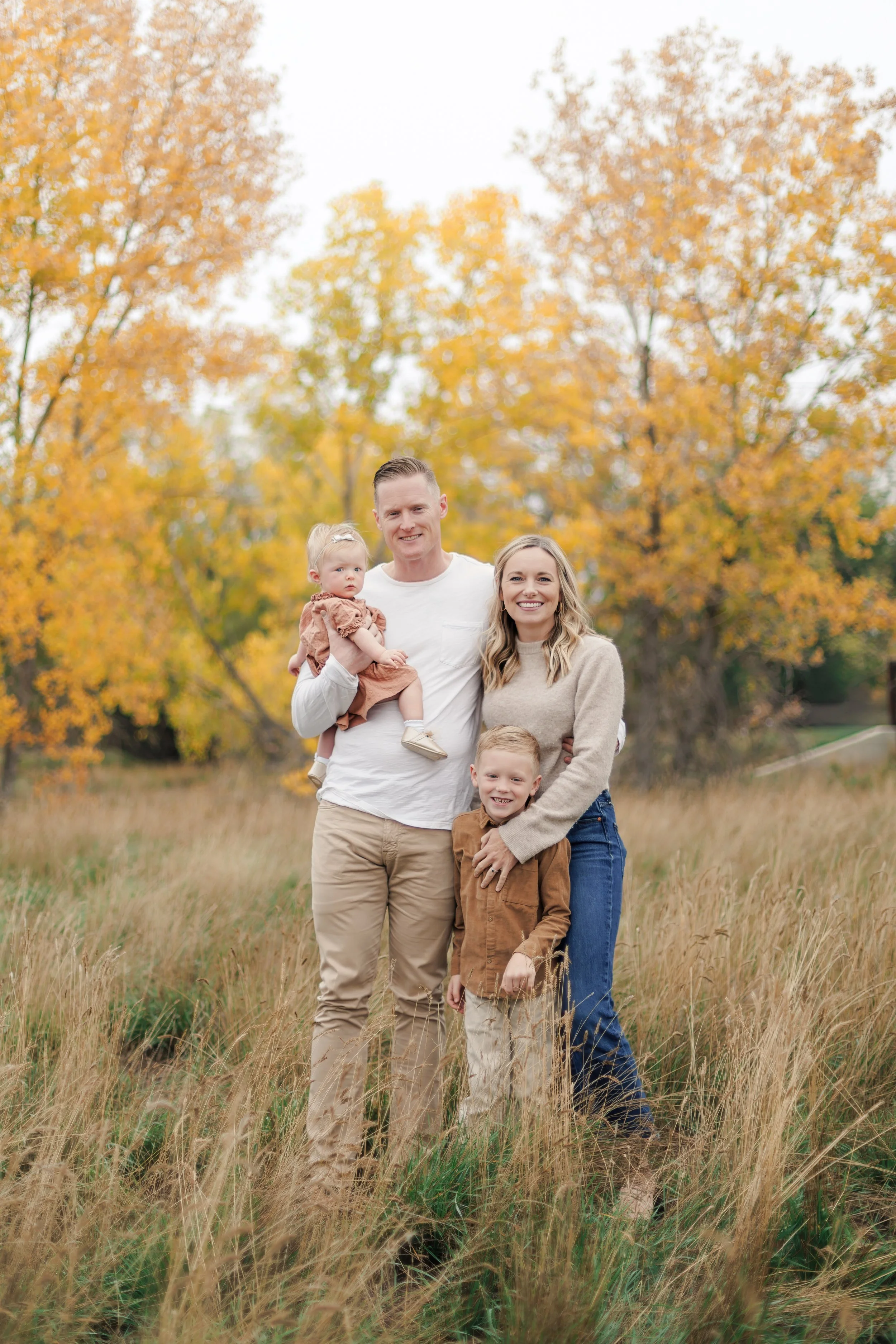 Family of four standing in a field with fall foliage in the background, smiling at the camera.