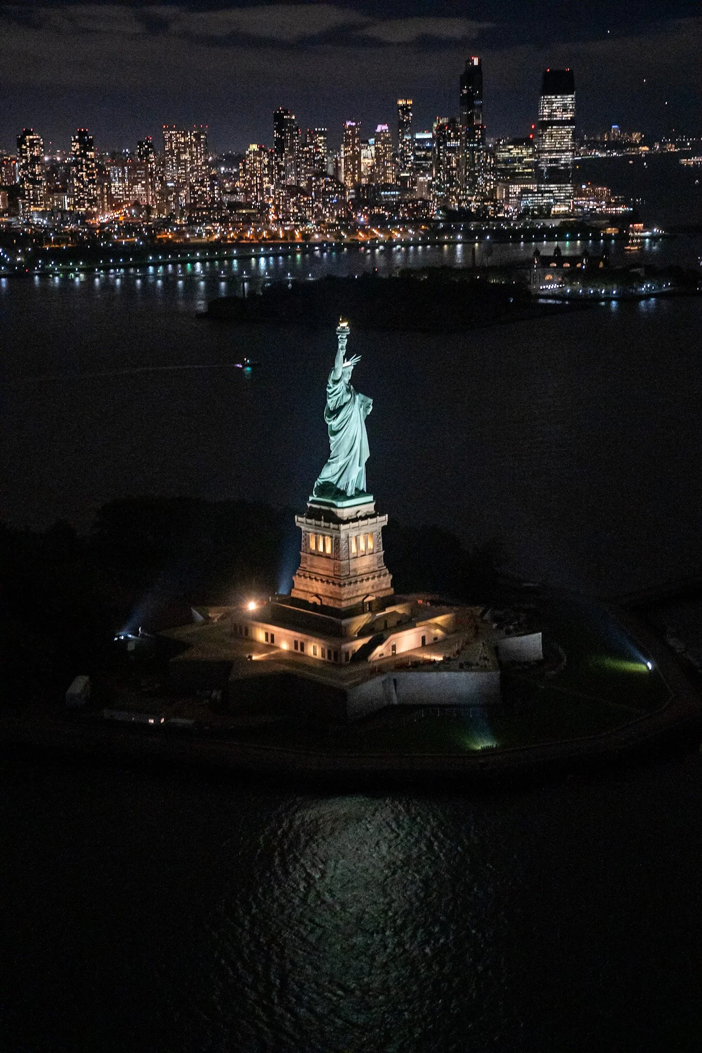Nighttime aerial view of the Statue of Liberty illuminated on Liberty Island with Manhattan skyline in the background.