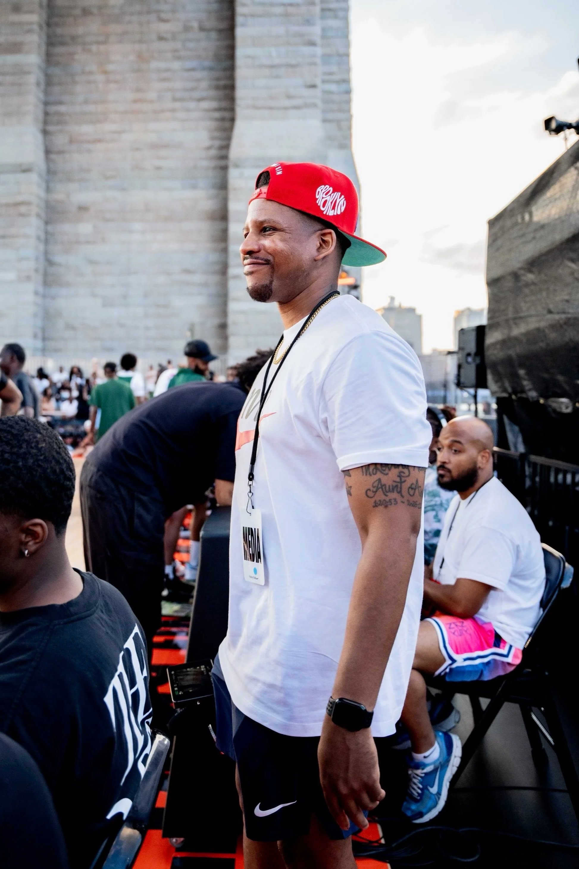 A man wearing a red baseball cap, white T-shirt, black Nike shorts, and a smartwatch standing at an outdoor event with others seated and standing around, large city buildings and a bridge in the background.