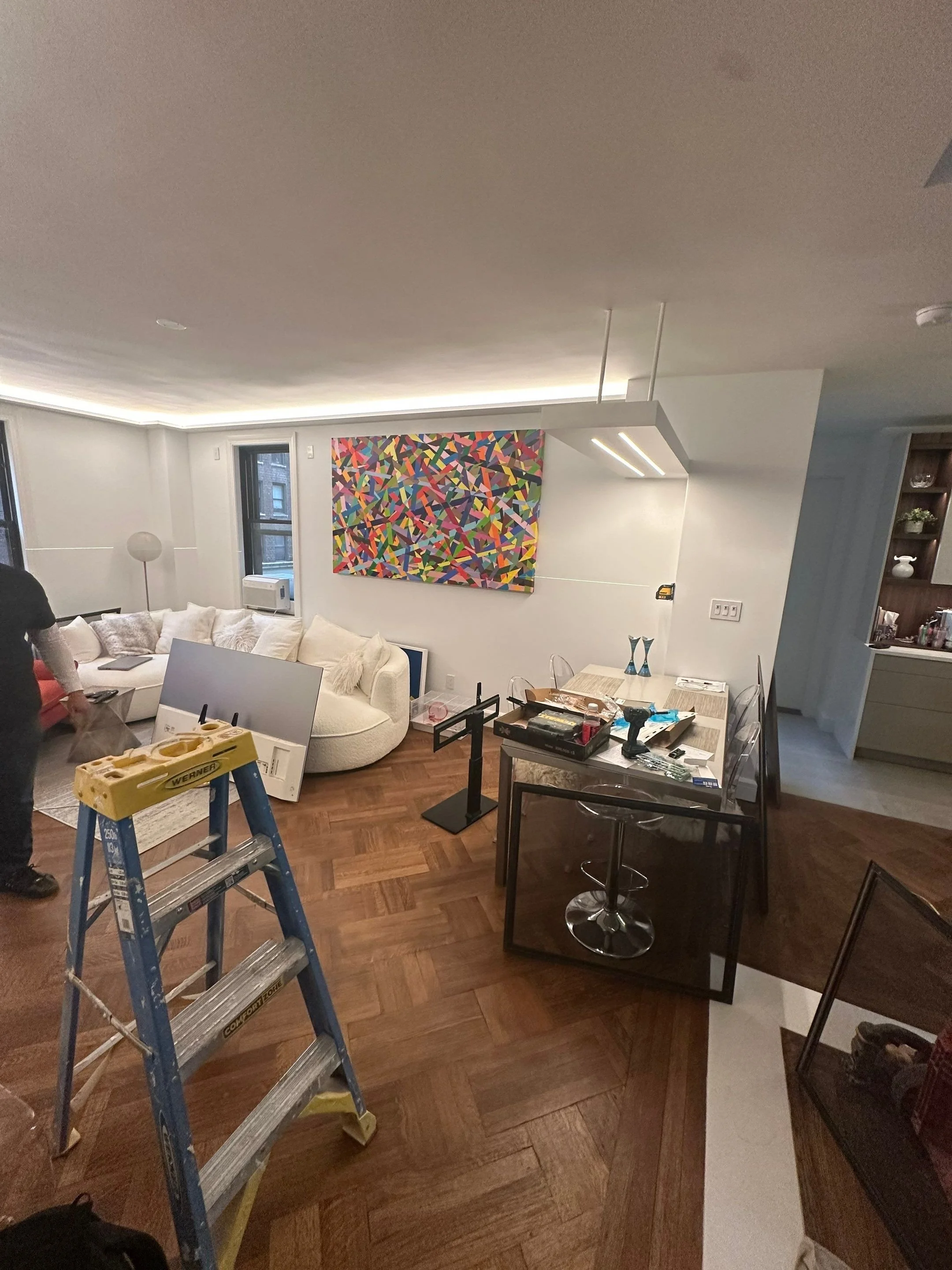 Living room with a colorful abstract painting on the wall, white sofa with pillows, wooden floor, ladder, and various furniture items being assembled or arranged.