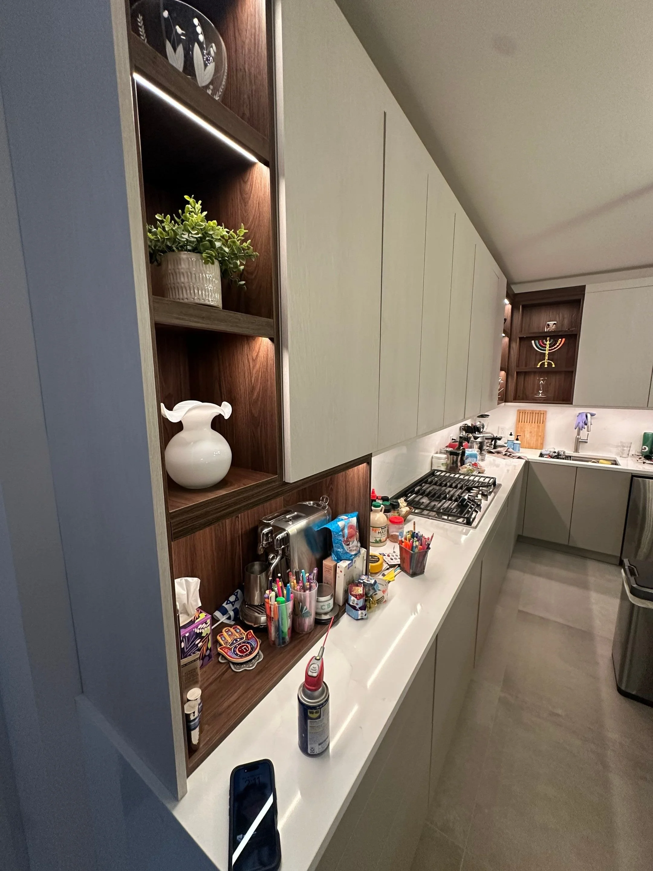 Messy kitchen countertop with various objects including pens, a lighter, and small containers, with kitchen cabinets and shelves in the background.