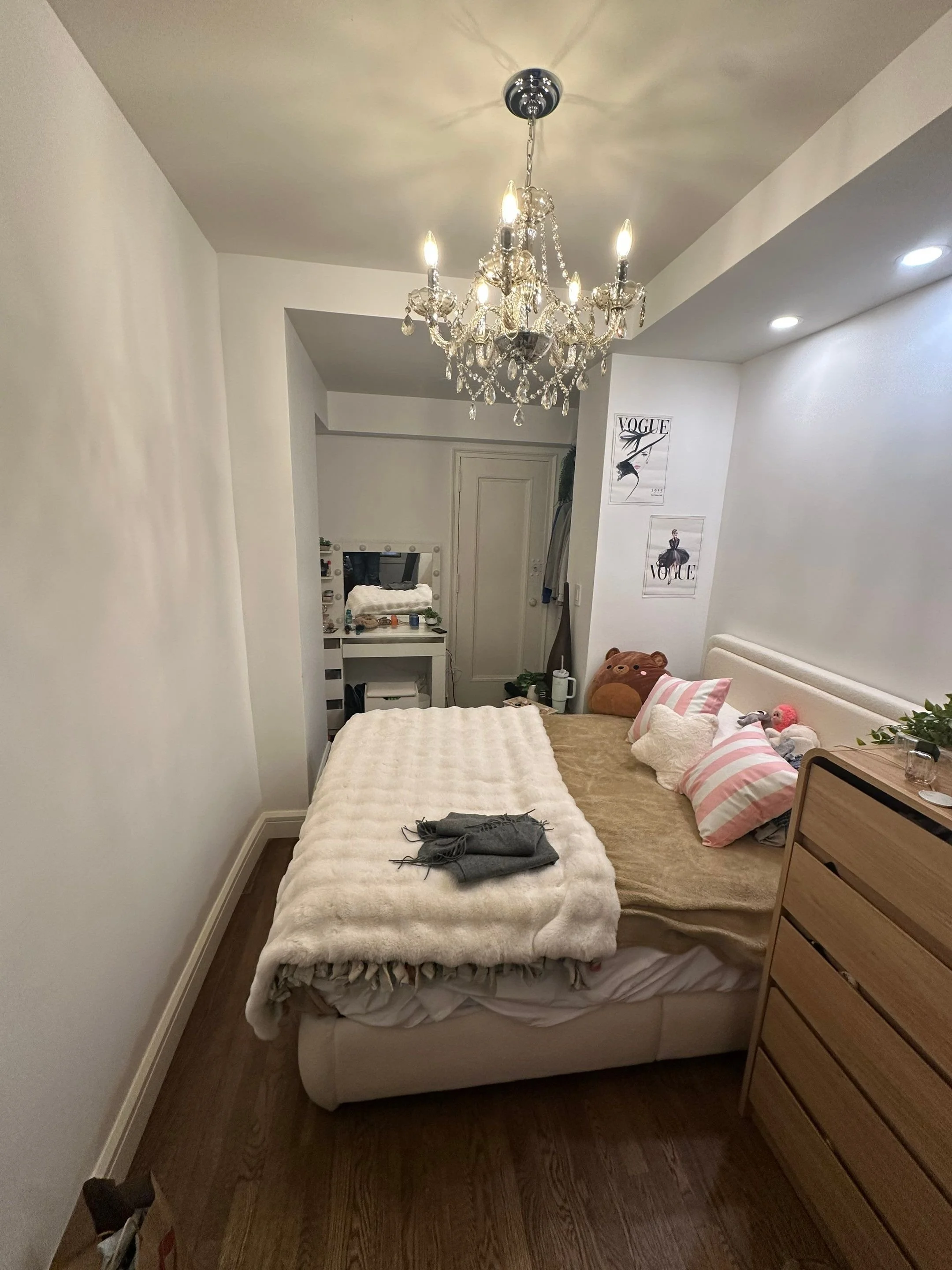 A bedroom with a bed, a chandelier overhead, and a wooden dresser. The bed has pink and white striped pillows and a large stuffed bear. There are pictures on the wall, a small vanity with mirror, and some clothing hanging on the wall.