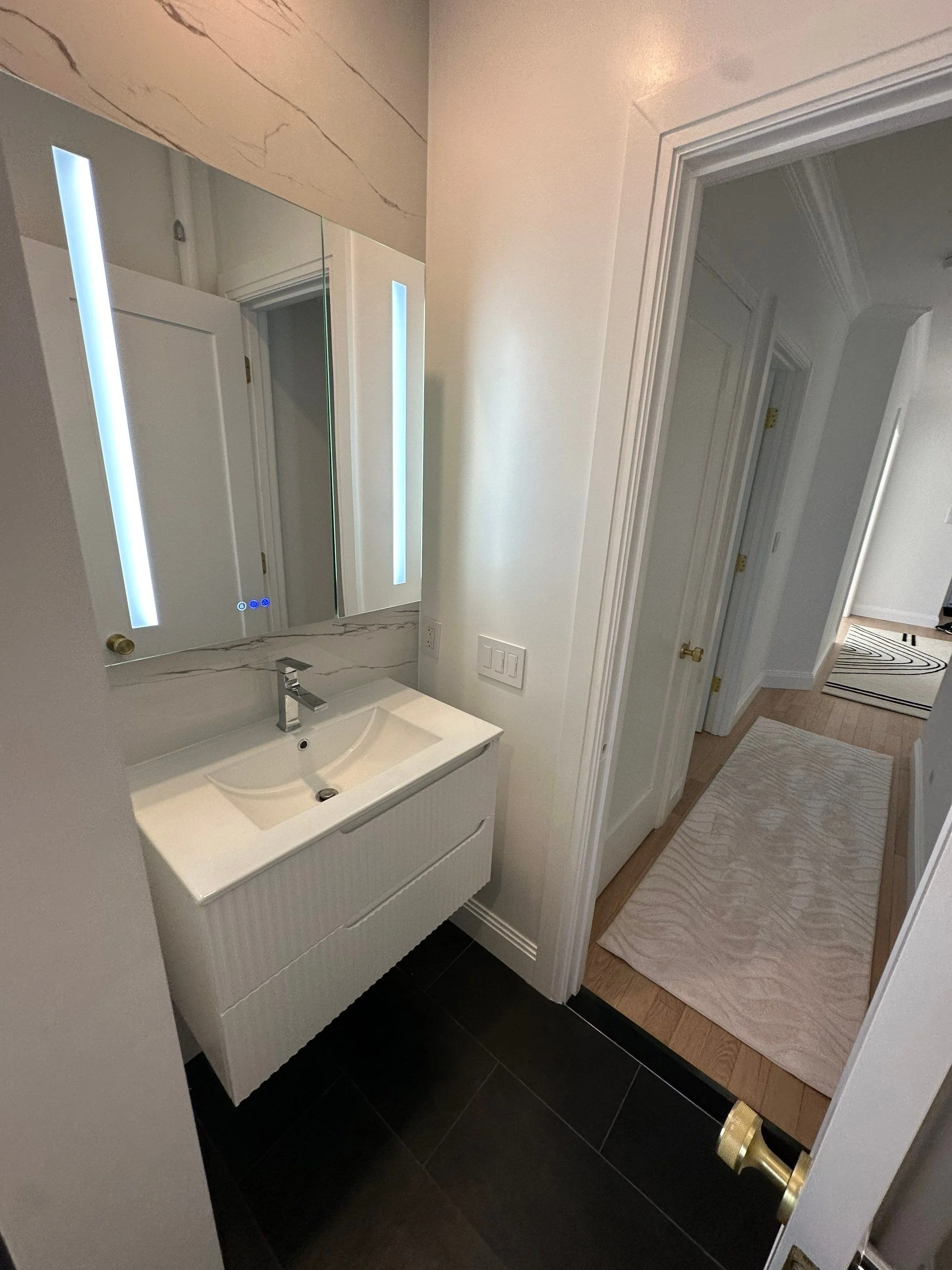 Bathroom vanity with a mirror and illuminated vertical light strips, black tiled floor, and a view into the hallway with a light-colored rug and wooden floors.
