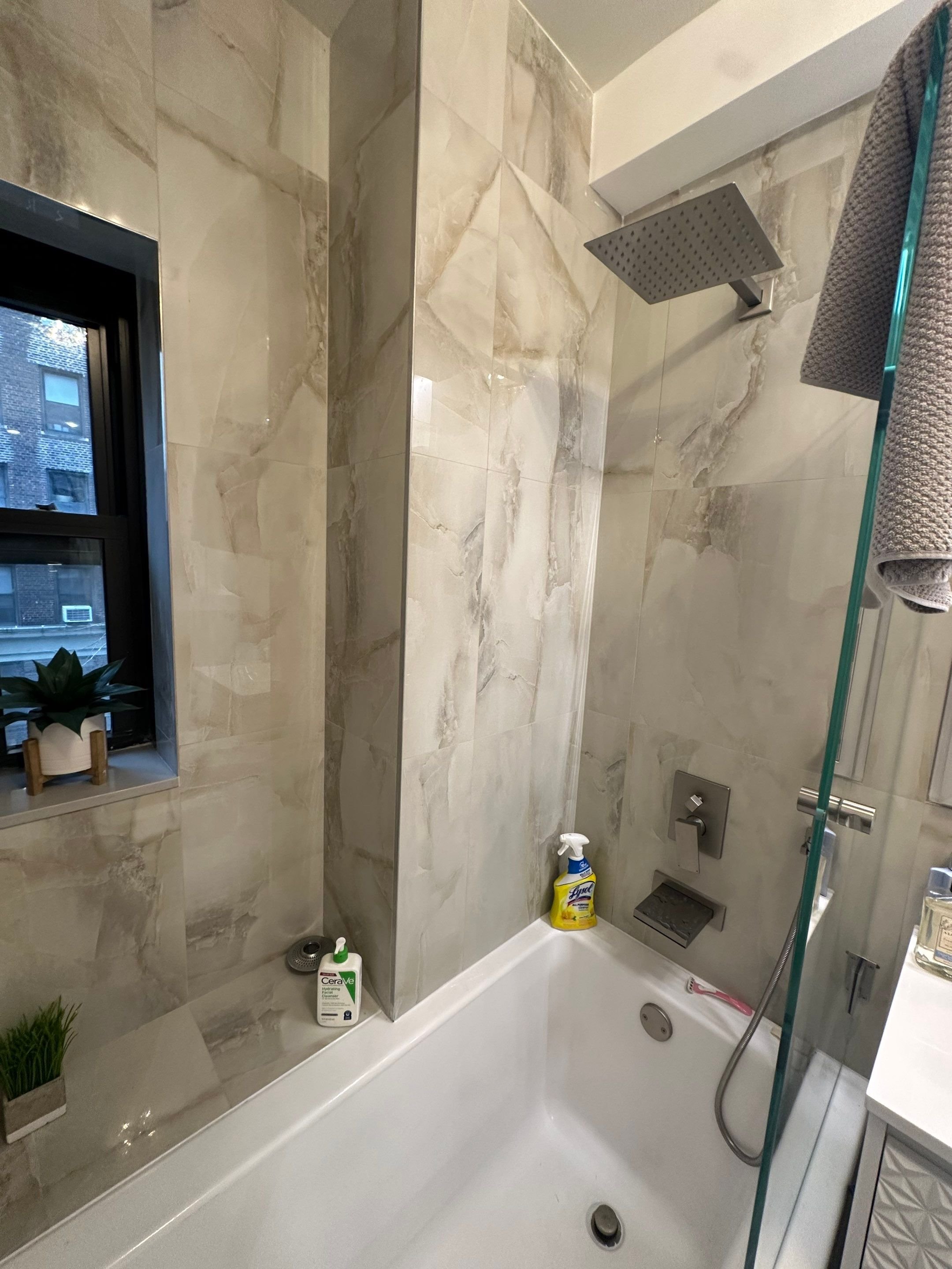 Modern bathroom shower area with marble walls, a rainfall showerhead, and a handheld shower. A small window with a potted plant, cleaning spray, and soap are visible.