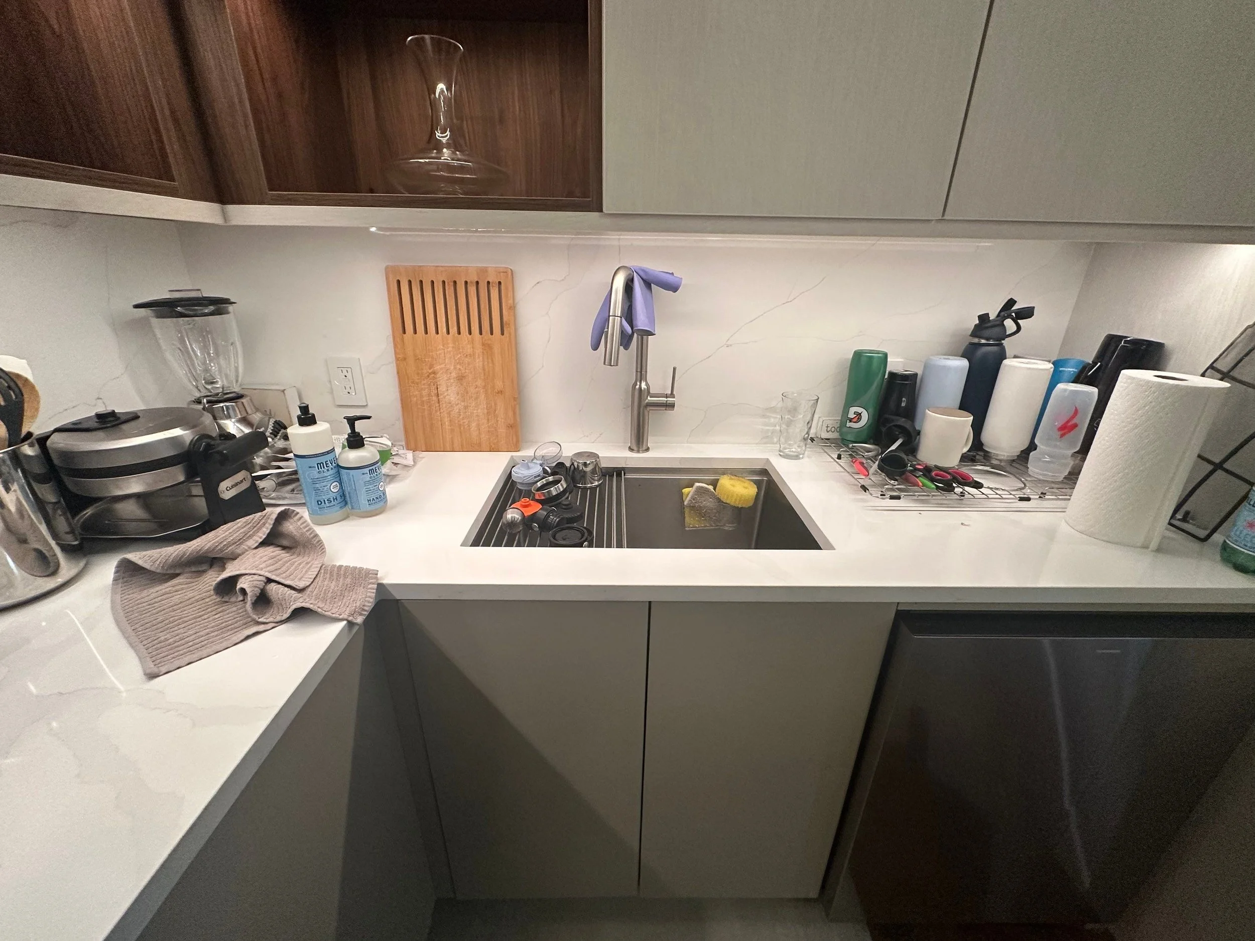 Modern kitchen counter with sink, dish soap, sponge, paper towels, appliances, utensils, and various drink containers.