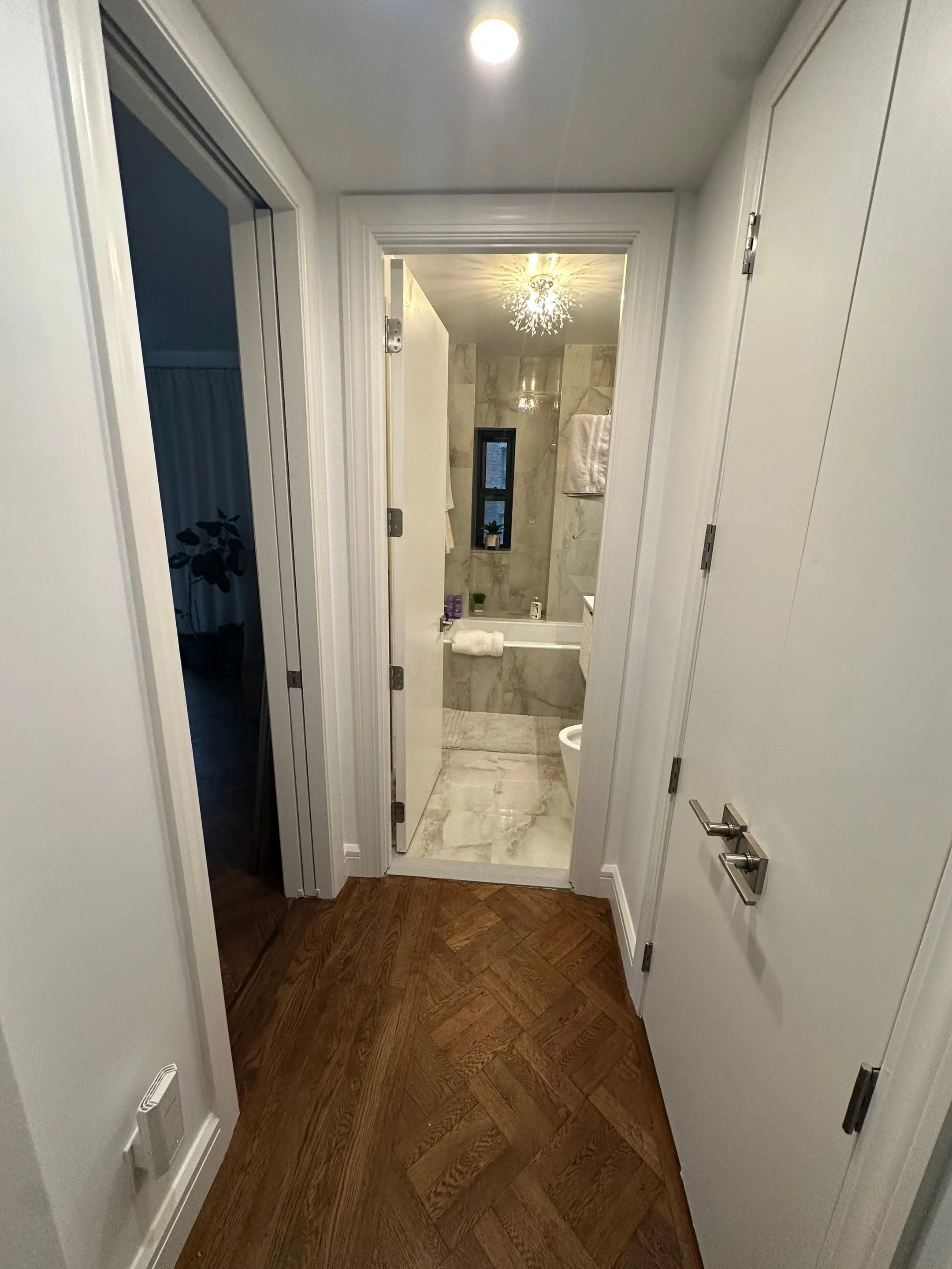 A view from a hallway looking into a bathroom with marble walls and floor. There is a small window, a towel rack with towels, and a ceiling light fixture. Part of a toilet is visible on the lower right.