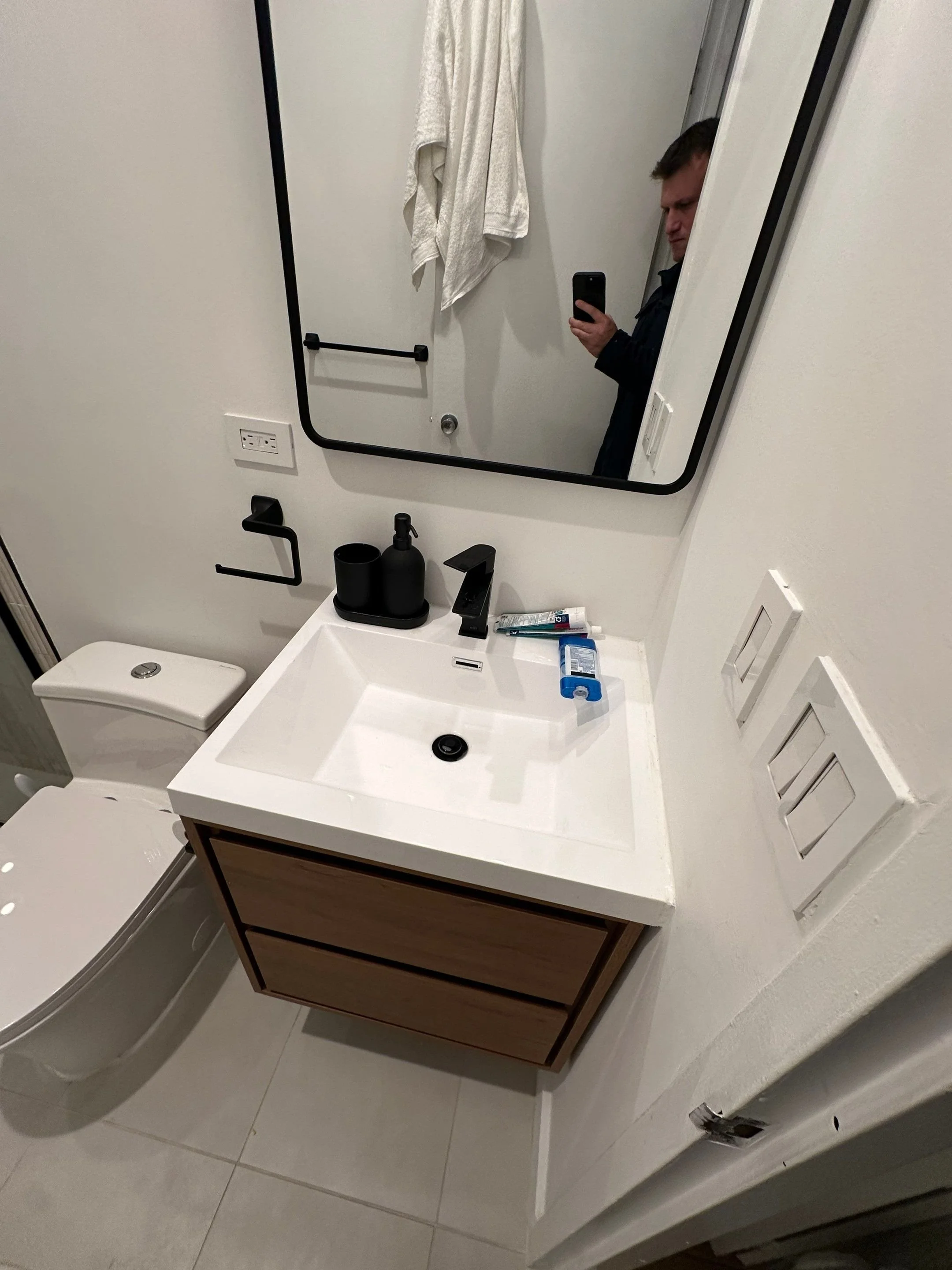 A bathroom with a white toilet, a vanity with a white sink, a mirror, and a man taking a selfie in the mirror. Bathroom accessories include a black soap dispenser, a cup, and toiletries on the sink. There are towels hanging on a wall hook and a towel bar.