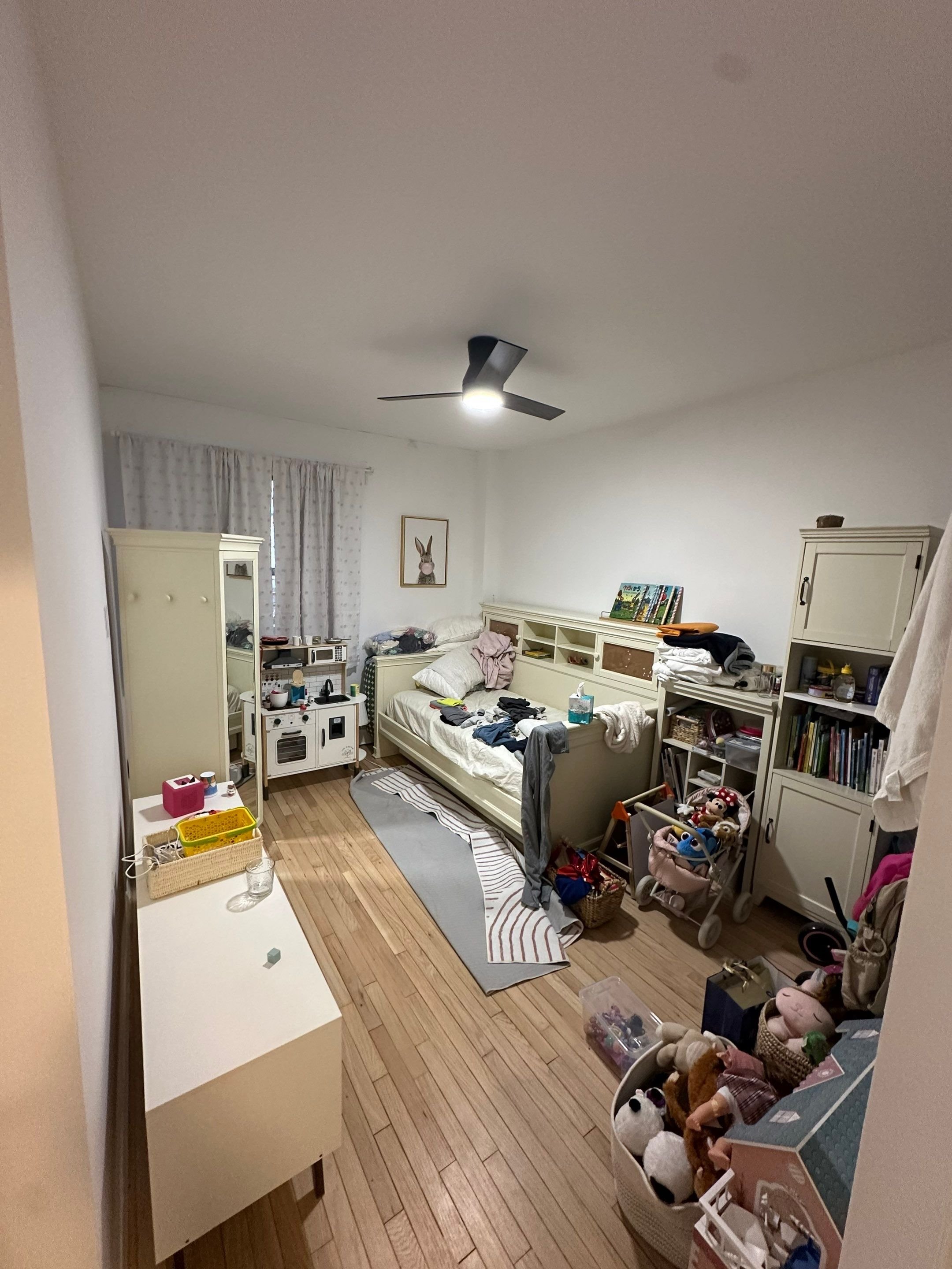 A messy bedroom with a bed in the center, clothes on it, and clutter around. Small furniture including a wardrobe, shelves, and a play kitchen. Stuffed animals and toys on the floor and in a basket. A ceiling fan is on the ceiling, and the room has white walls, a window with curtains, and a hardwood floor.