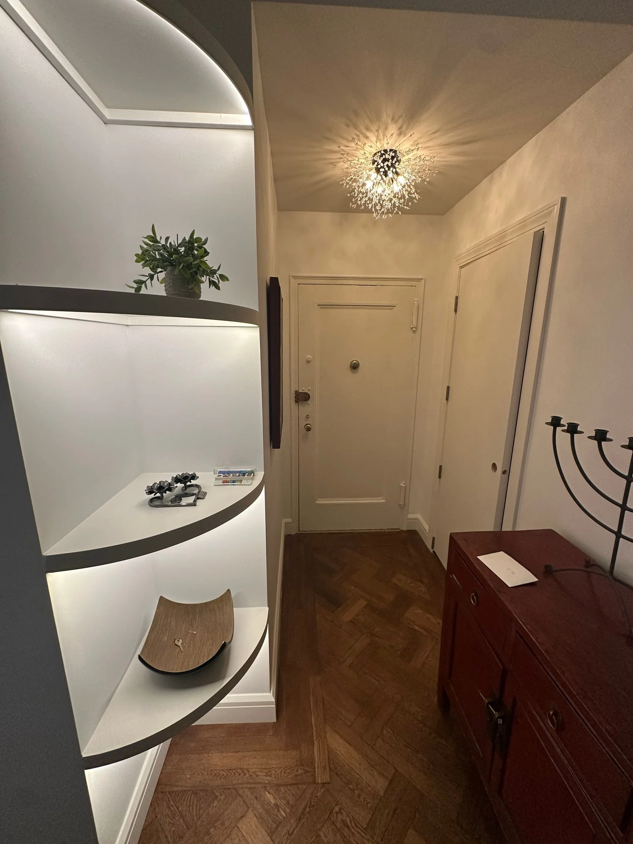 View of an entryway with a white illuminated wall shelf holding a potted plant, keys, and small items. A decorative chandelier with numerous branches and lights is on the ceiling. The front door is white with a doorbell and peephole. To the right, a red wooden cabinet with a candelabrum on top is partially visible.