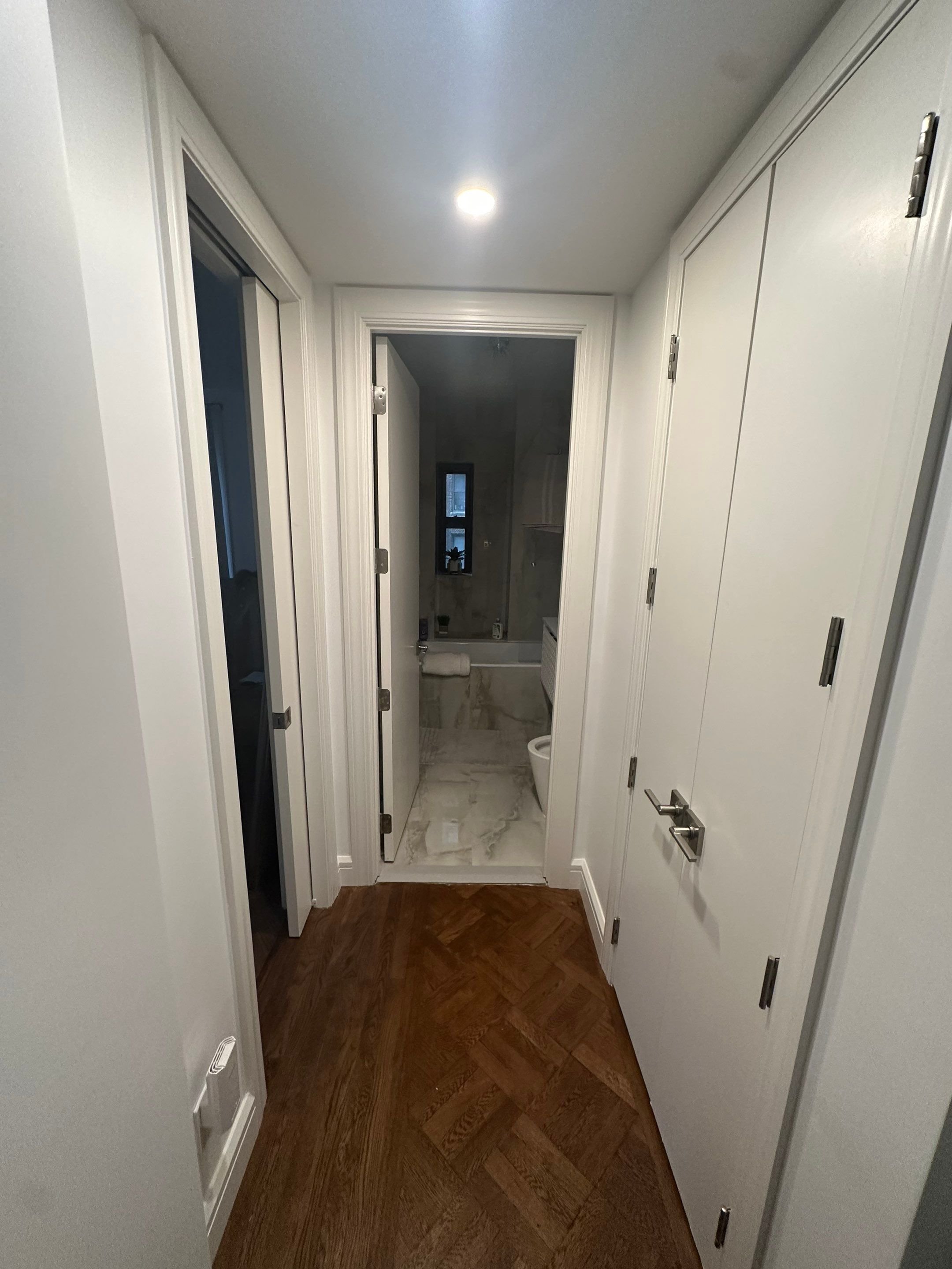 A hallway leading to a bathroom with hardwood flooring and white walls. The bathroom has a marble floor and is lit by a ceiling light, with a small window and visible fixtures.