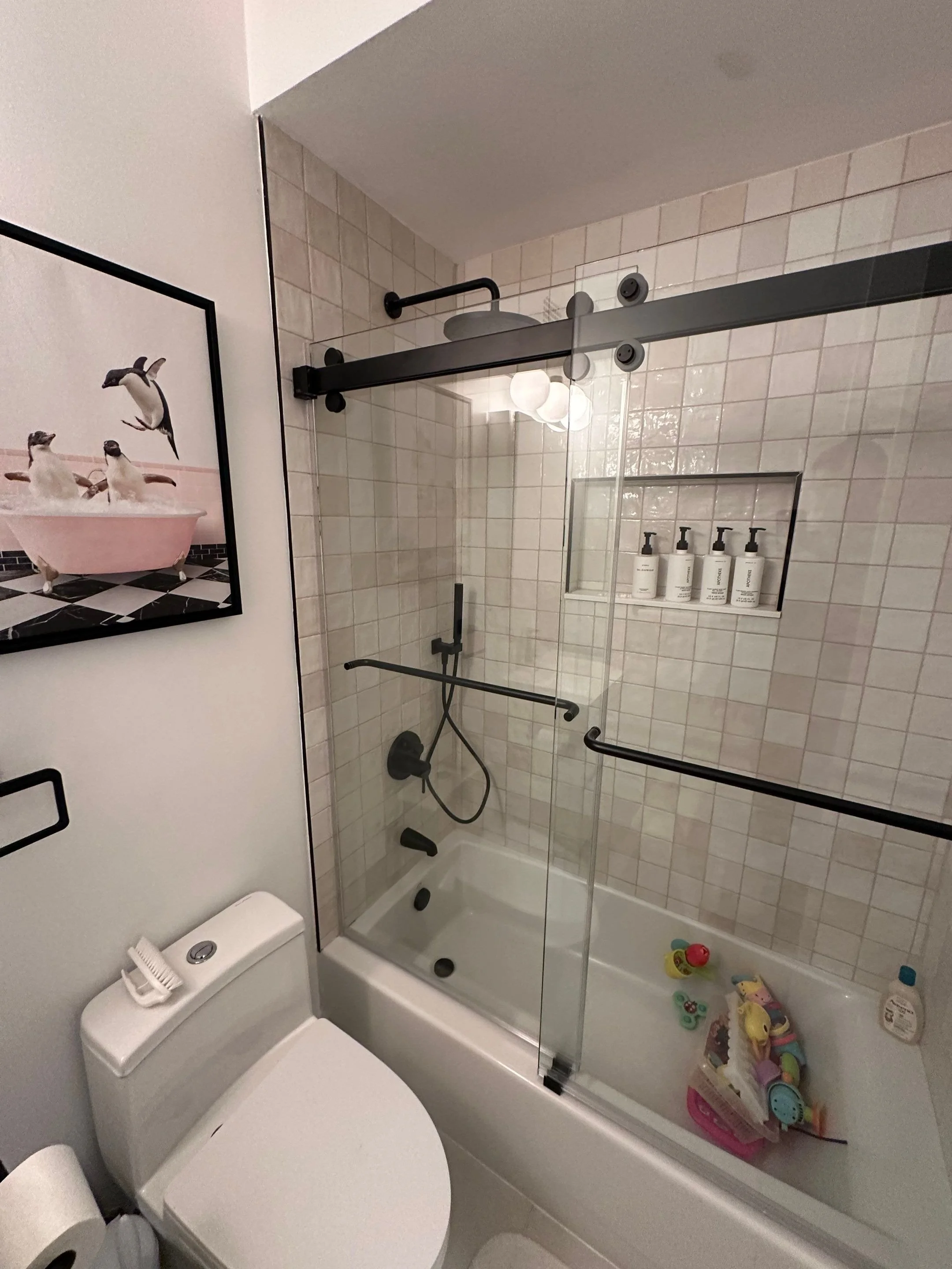 Bathroom with a shower and bathtub combination, a toilet, a framed picture of penguins, a soap container on the bathtub, and toys in the bathtub. The shower has a sliding glass door with black hardware and white tiled walls.