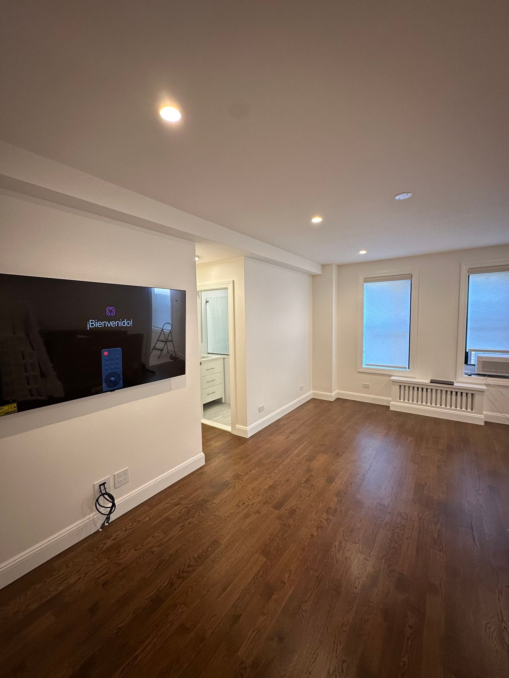 Empty living room with hardwood floors, white walls, and three windows with blinds, featuring a wall-mounted TV displaying a welcome message in Spanish, and a small hallway leading to a bathroom.