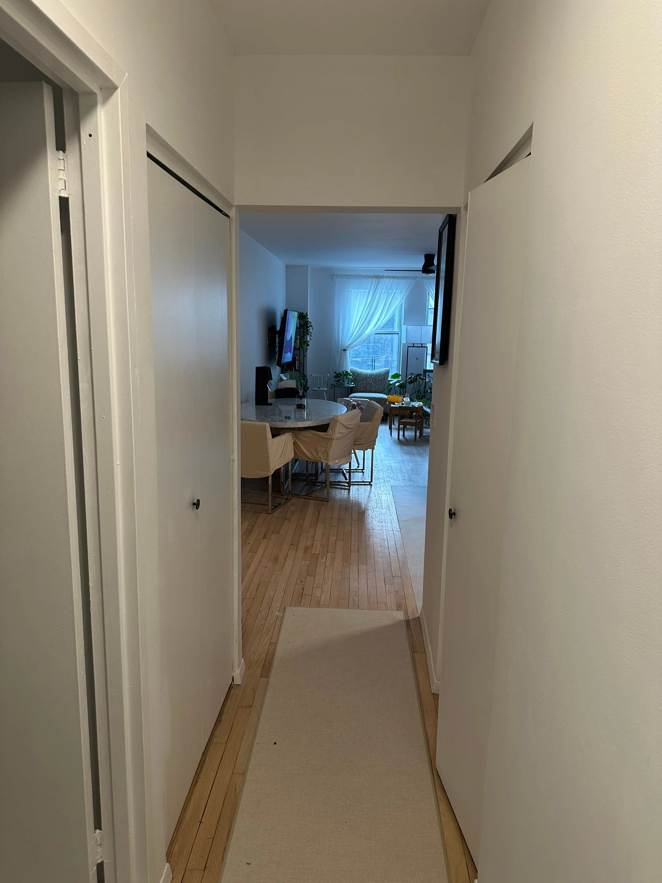 A hallway view looking into a living room with a dining table, chairs, and a couch by a window with curtains.