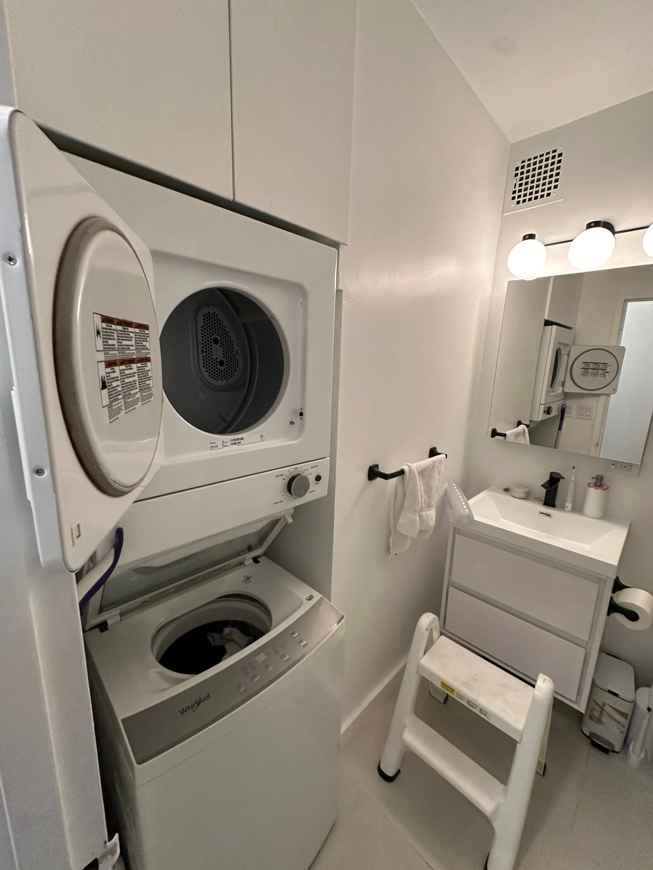 Laundry room with stacked washer and dryer, bathroom vanity with mirror and light fixtures, towel bar with a towel, small step ladder, and various bathroom accessories.
